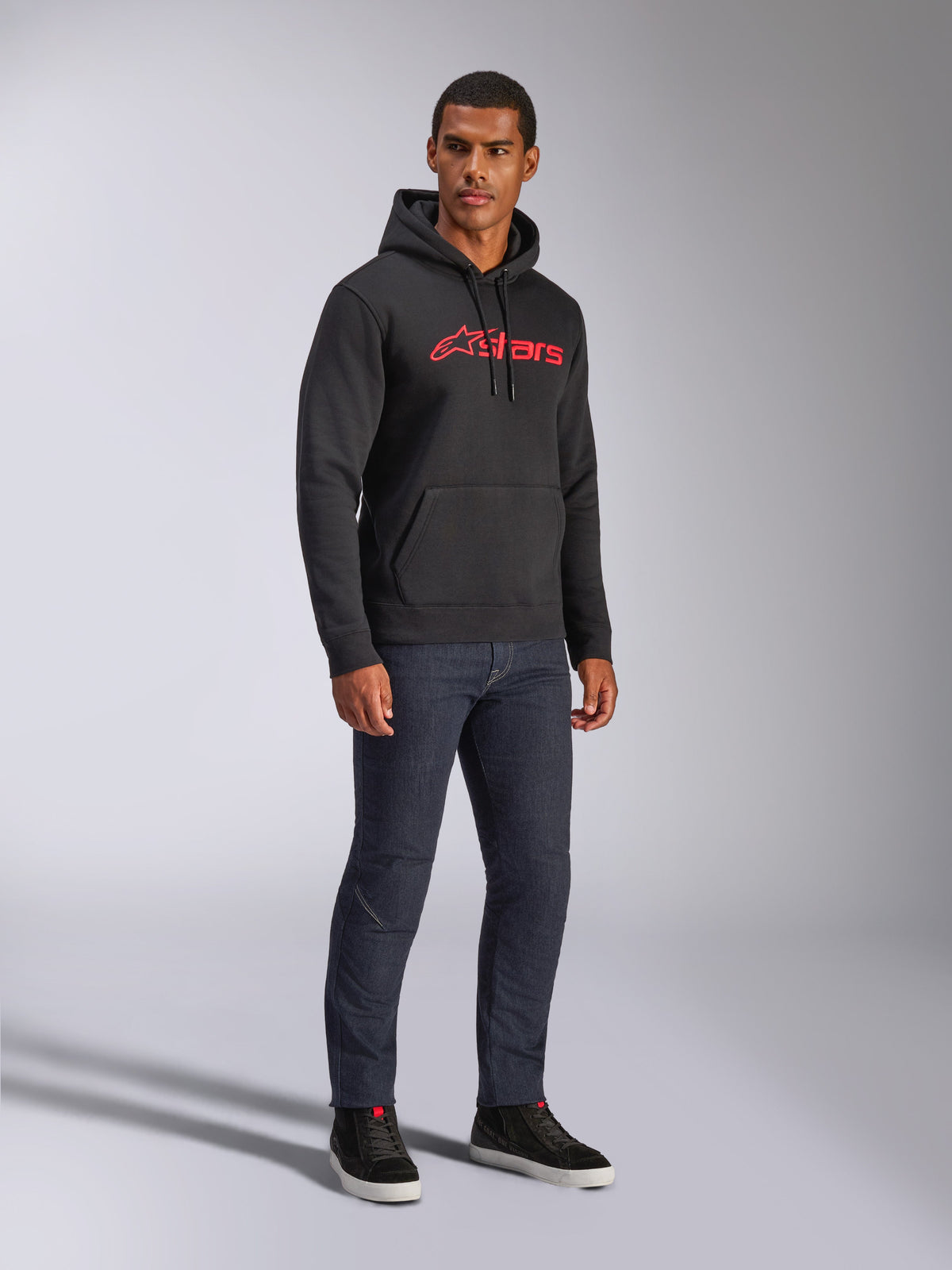 Alpinestars Blaze V3 Hoodie, schwarzer Fleece-Kapuzenpullover mit rotem Alpinestars-Logo auf der Brust, getragen von einem männlichen Model mit dunklen Denim-Jeans und schwarzen Sneakern, lässige Streetwear im Motorsport-Stil