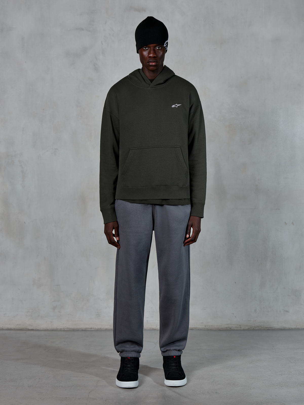 Model trägt Alpinestars Essentials Ageless Hoodie, lässige Streetwear, Off Black mit kleinem weißem Logo auf der Brust, kombiniert mit grauer Jogginghose, schwarzer Beanie und schwarzen Sneakern, minimalistisches, vom Motorsport inspiriertes Design