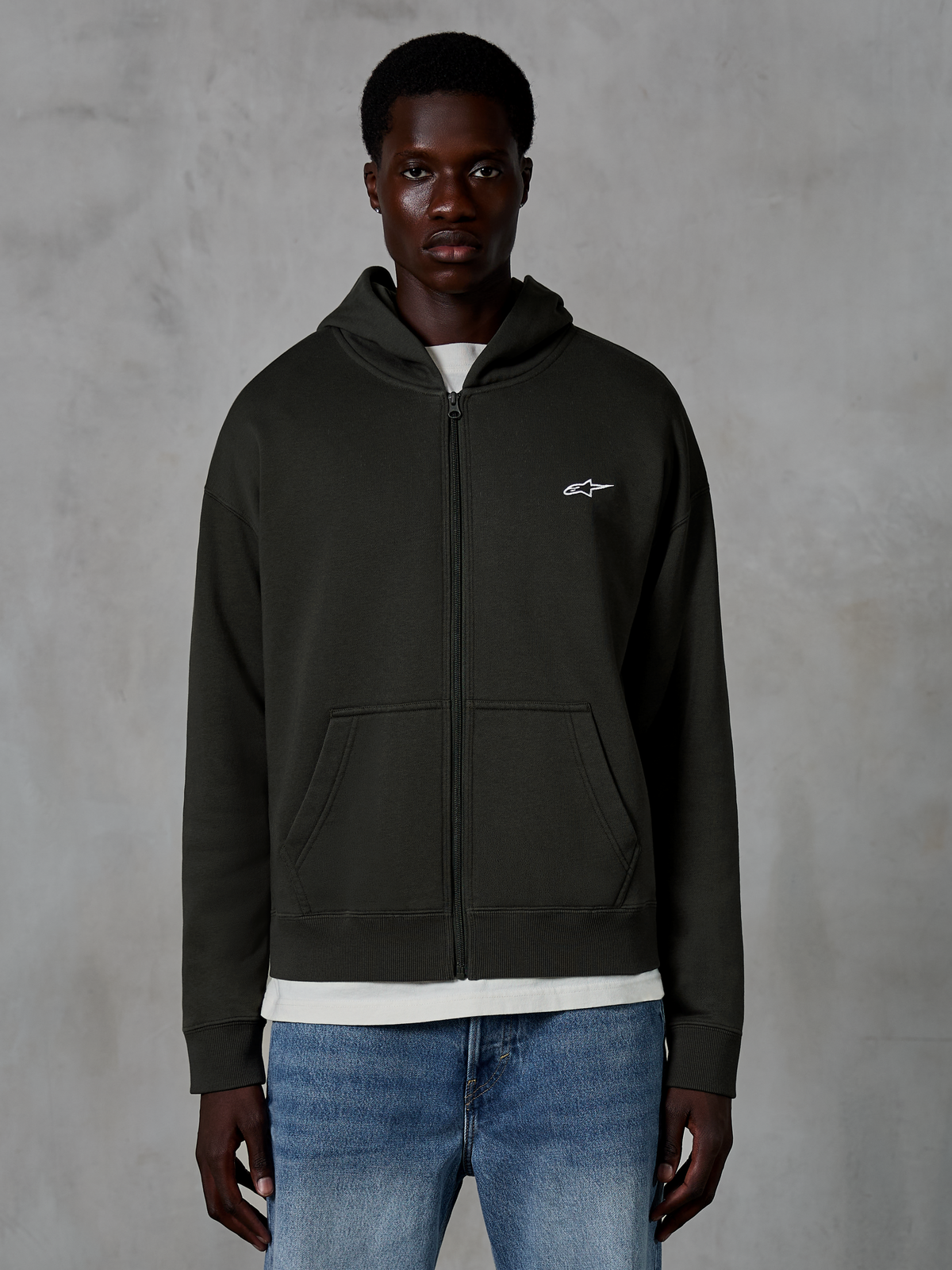Alpinestars Essentials Ageless Zip Hoodie, Full-Zip Hoodie, Off Black, featuring a small white star logo on the chest, front pockets, and ribbed cuffs, worn as casual motorsport-inspired streetwear