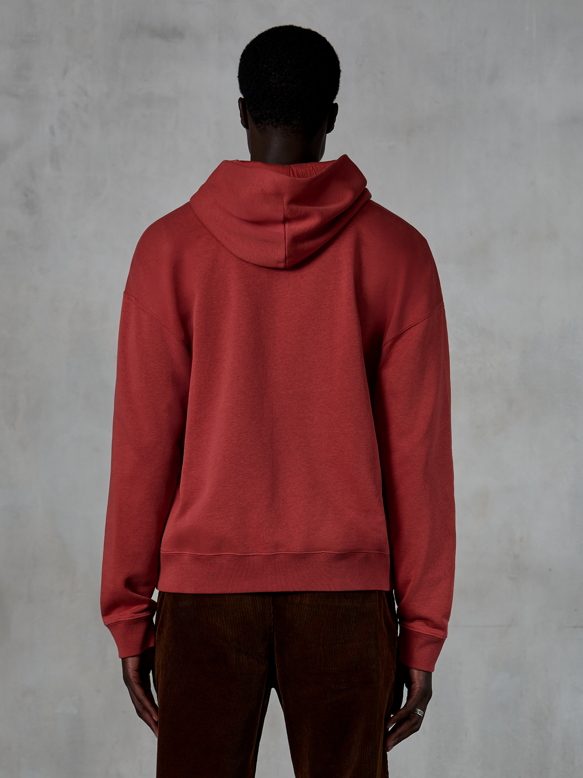 Alpinestars Essentials Ageless Zip Hoodie, Ziegelrot, Rückansicht zeigt entspannte Passform und Kapuze mit Kordelzug, kombiniert mit brauner Cordhose, lässige, vom Motorsport inspirierte Streetwear