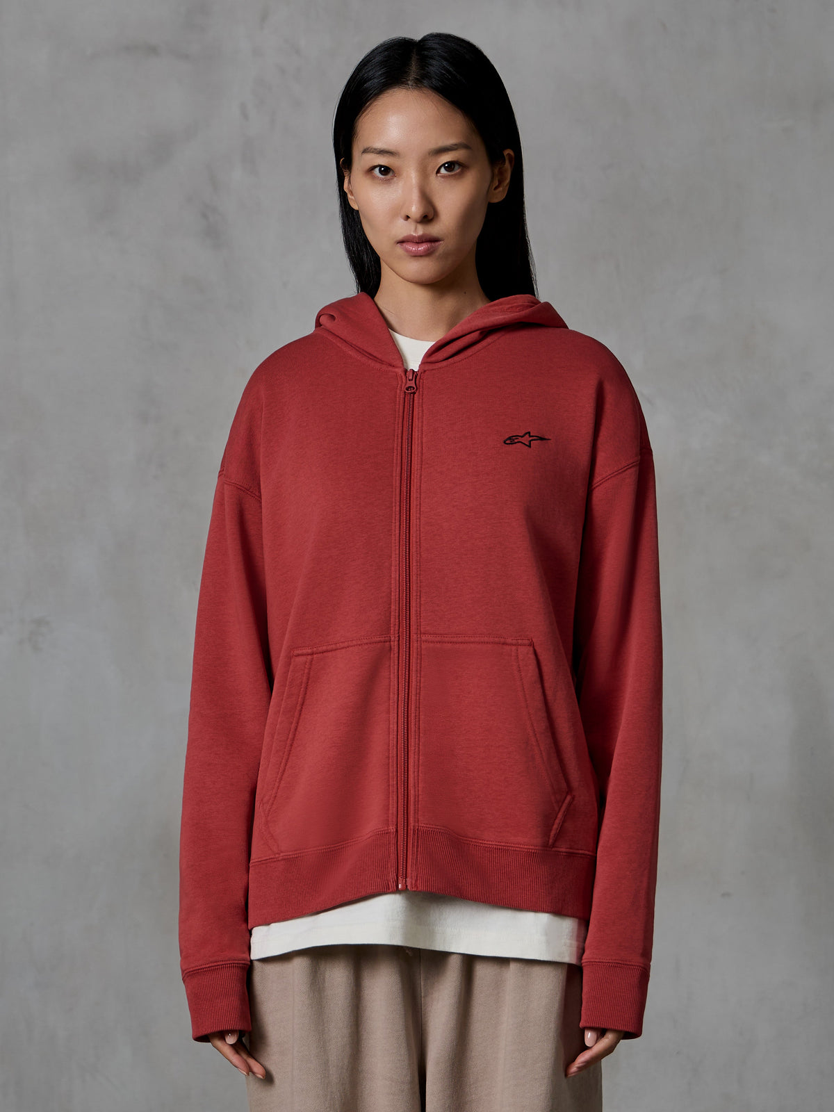 Alpinestars Essentials Ageless Zip Hoodie, Ziegelrot, lässige Streetwear, Modell trägt Hoodie mit durchgehendem Reißverschluss und Kapuze, kleines schwarzes Alpinestars-Logo auf der Brust, entspannte Passform, Kompakt-Fleece.