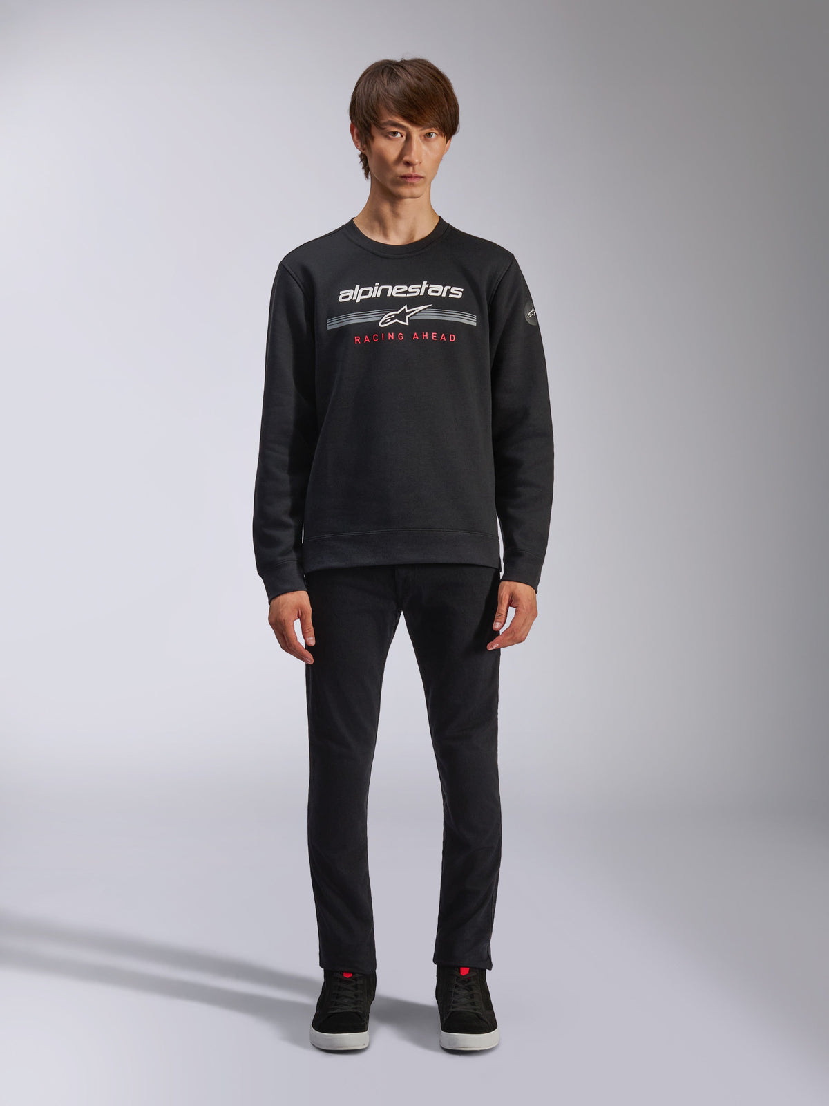 Alpinestars Bettern Crew, schwarzes Sweatshirt mit Rundhalsausschnitt, zeigt weißes Alpinestars-Logo mit grauen horizontalen Streifen und rotem „RACING AHEAD“-Text, lässige Motorsport-Streetwear, getragen von einem Model mit schwarzer Hose und Sneakern