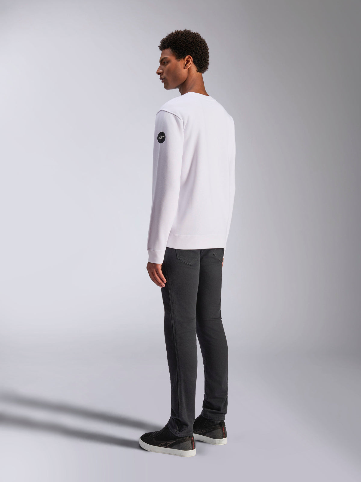 Alpinestars Bettern Crew, weißes Crewneck-Sweatshirt, Rück- und Seitenansicht, schwarzes kreisförmiges Logo am Ärmel, getragen mit dunkelgrauer Hose und schwarzen Sneakern, lässige Motorsport-Lifestyle-Streetwear
