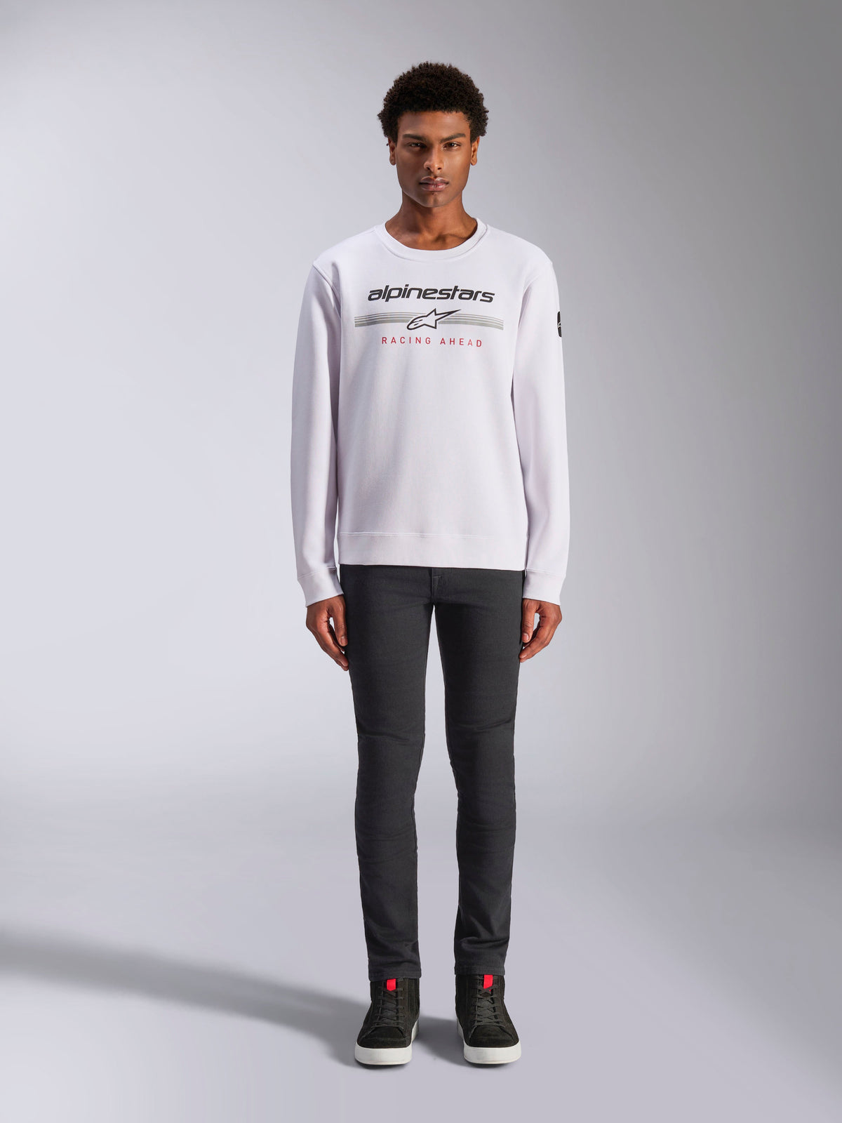 Alpinestars Bettern Crew, weißes Sweatshirt mit Rundhalsausschnitt, getragen von einem männlichen Model mit dunkelgrauer Hose und schwarzen High-Top-Sneakern, Alpinestars-Branding mit rotem 'Racing Ahead'-Schriftzug, lässige, vom Motorsport inspirierte Streetwear