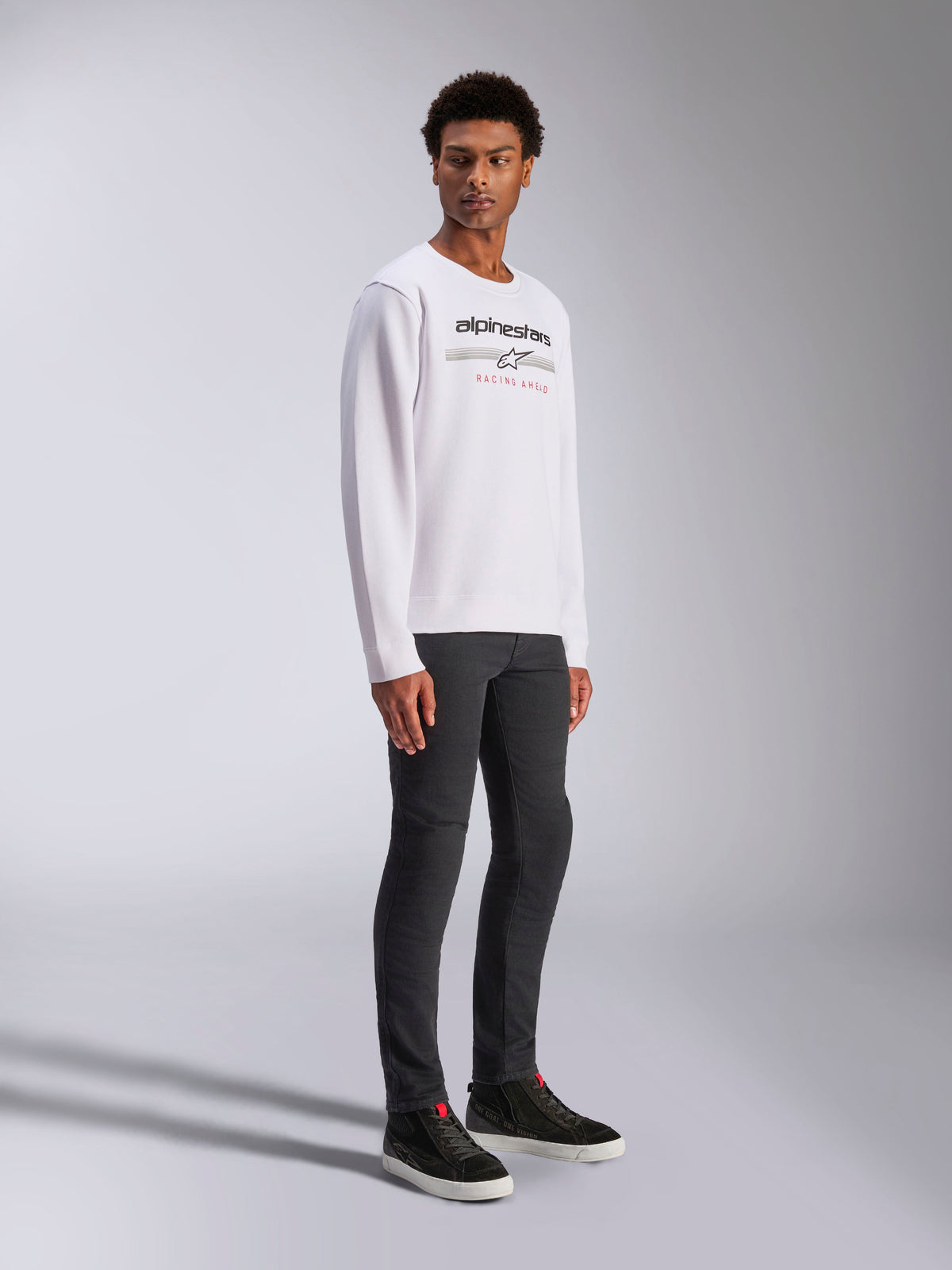 Alpinestars Bettern Crew, weißes Pullover-Sweatshirt mit schwarzem Alpinestars-Logo und rotem „Racing Ahead“-Schriftzug, lässige Motorsport-Streetwear, getragen von einem Model mit dunkler Hose und schwarzen Sneakern