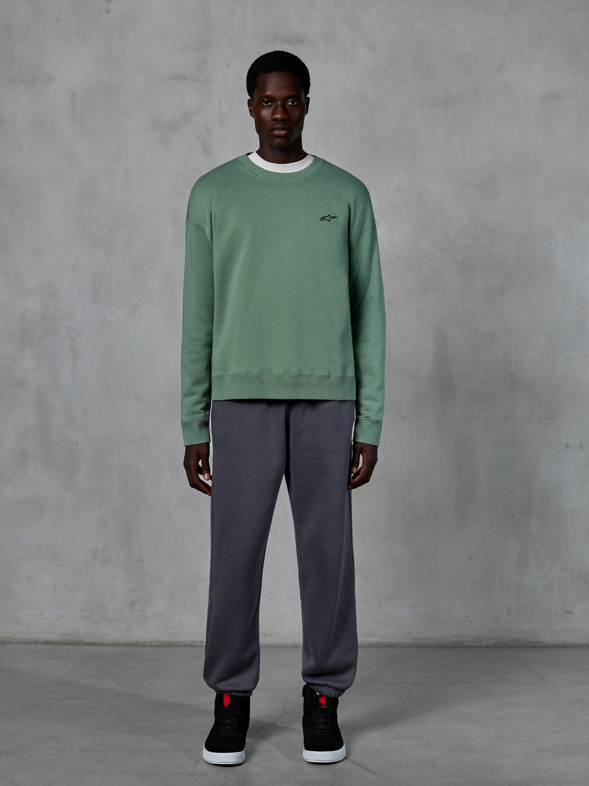 Alpinestars Essentials Ageless Crew, Rundhals-Sweatshirt in Cactus-Grün, mit kleinem schwarzem Logo auf der Brust, getragen von einem Model mit grauer Jogginghose und schwarzen Sneakern, lässige vom Motorsport inspirierte Streetwear