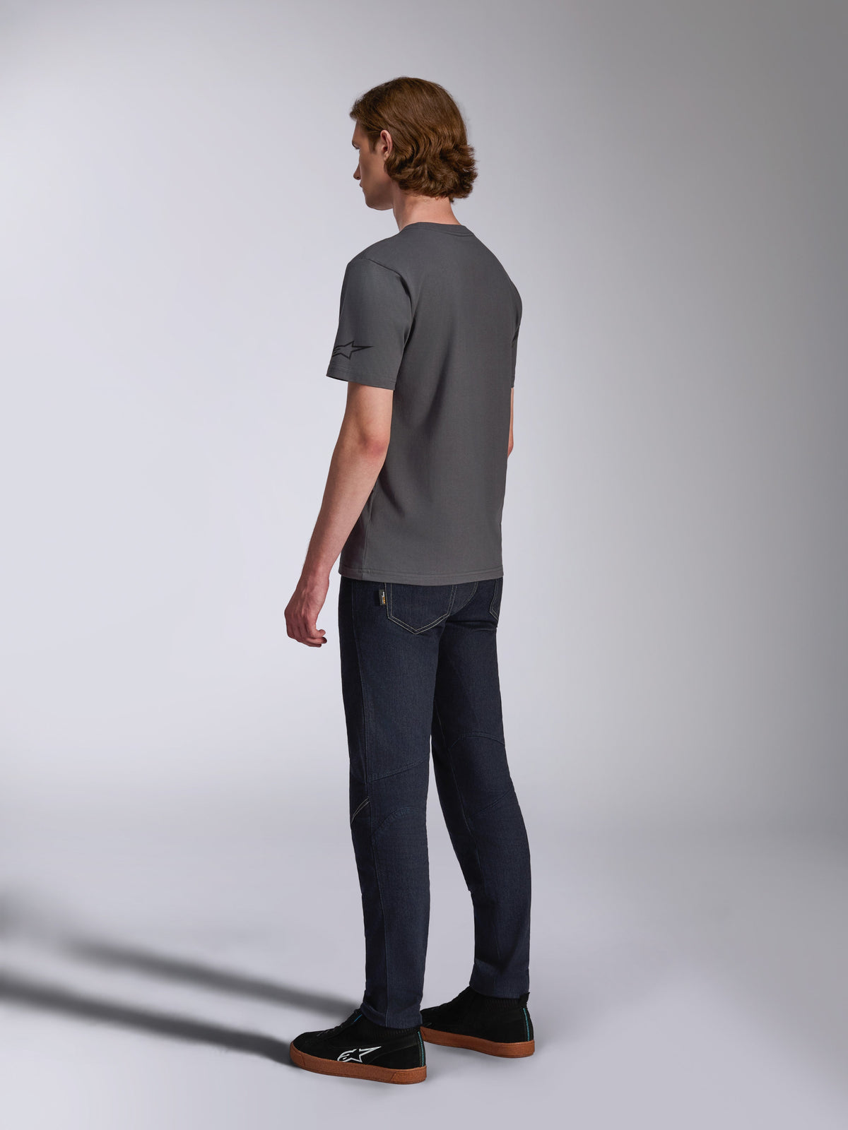 Alpinestars Waving CSF Tee, Kurzarm-T-Shirt, Anthrazit, Rückansicht am Model, kleines schwarzes Stern-Logo am Ärmel, getragen mit dunklen Denim-Jeans und schwarzen Sneakern, lässige Motorsport-Streetwear