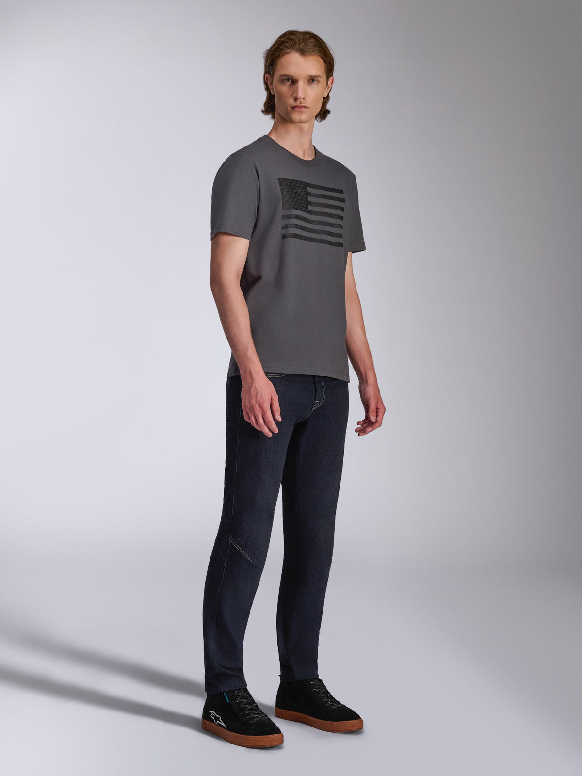 Alpinestars Waving CSF Tee, Kurzarm-T-Shirt, Charcoal, Grafik mit schwarzer wehender Flagge, getragen von einem Model mit dunkler Jeans und Sneakern, Casual Motorsport Streetwear