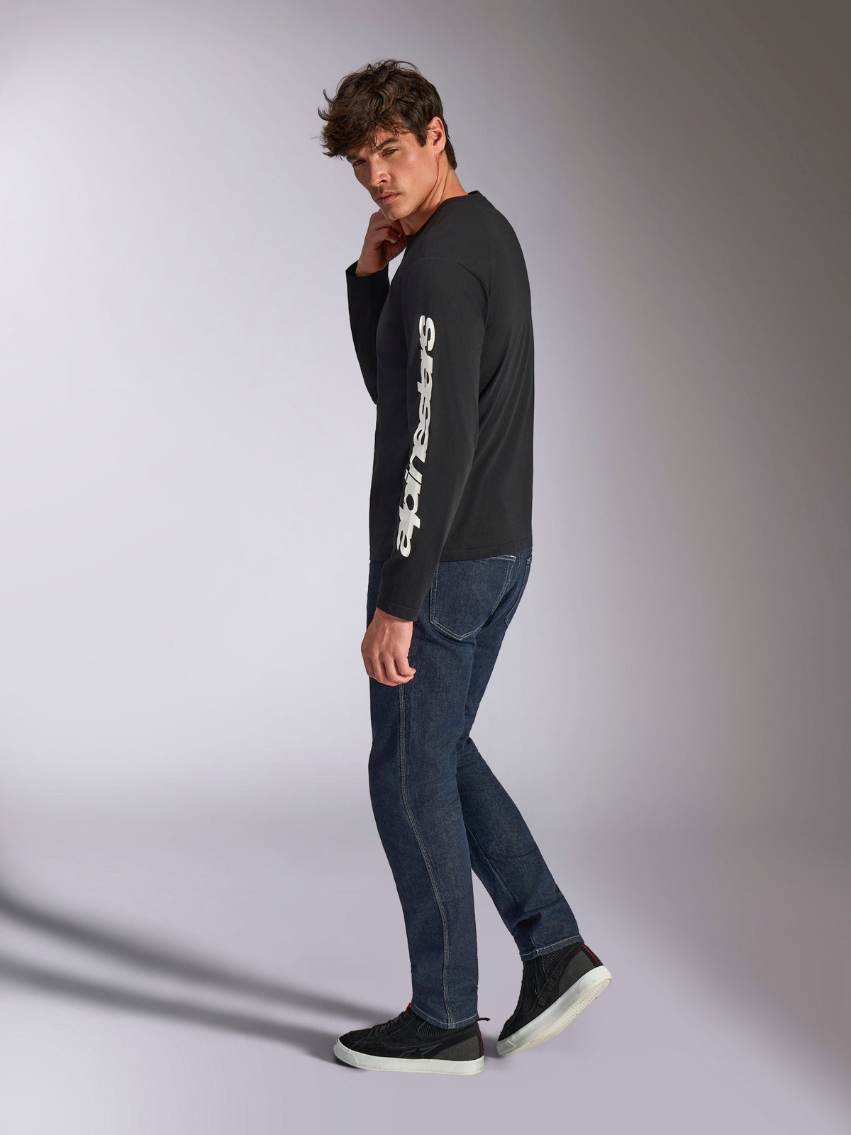Alpinestars Trackside CSF Long Sleeve Tee, Black with white vertical Alpinestars wordmark on the sleeve, worn by a model with dark denim and sneakers, casual motorsport-inspired sportswear