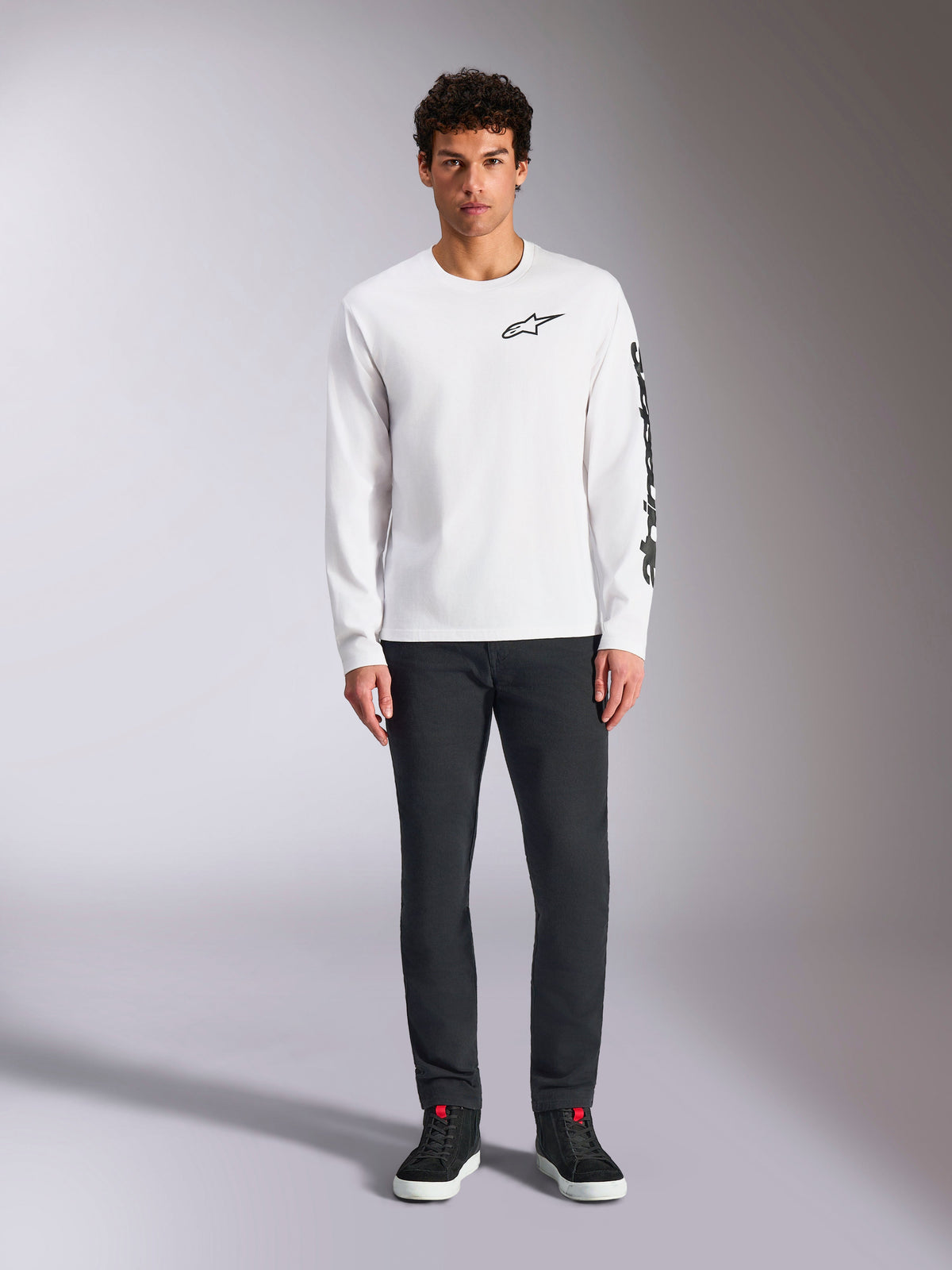 Alpinestars Trackside CSF Langarmshirt, Weiß mit schwarzem Branding, kleines Logo auf der Brust und vertikaler Schriftzug am Ärmel, getragen von einem Model mit dunkler Hose und schwarzen Sneakern, lässige, vom Motorsport inspirierte Streetwear