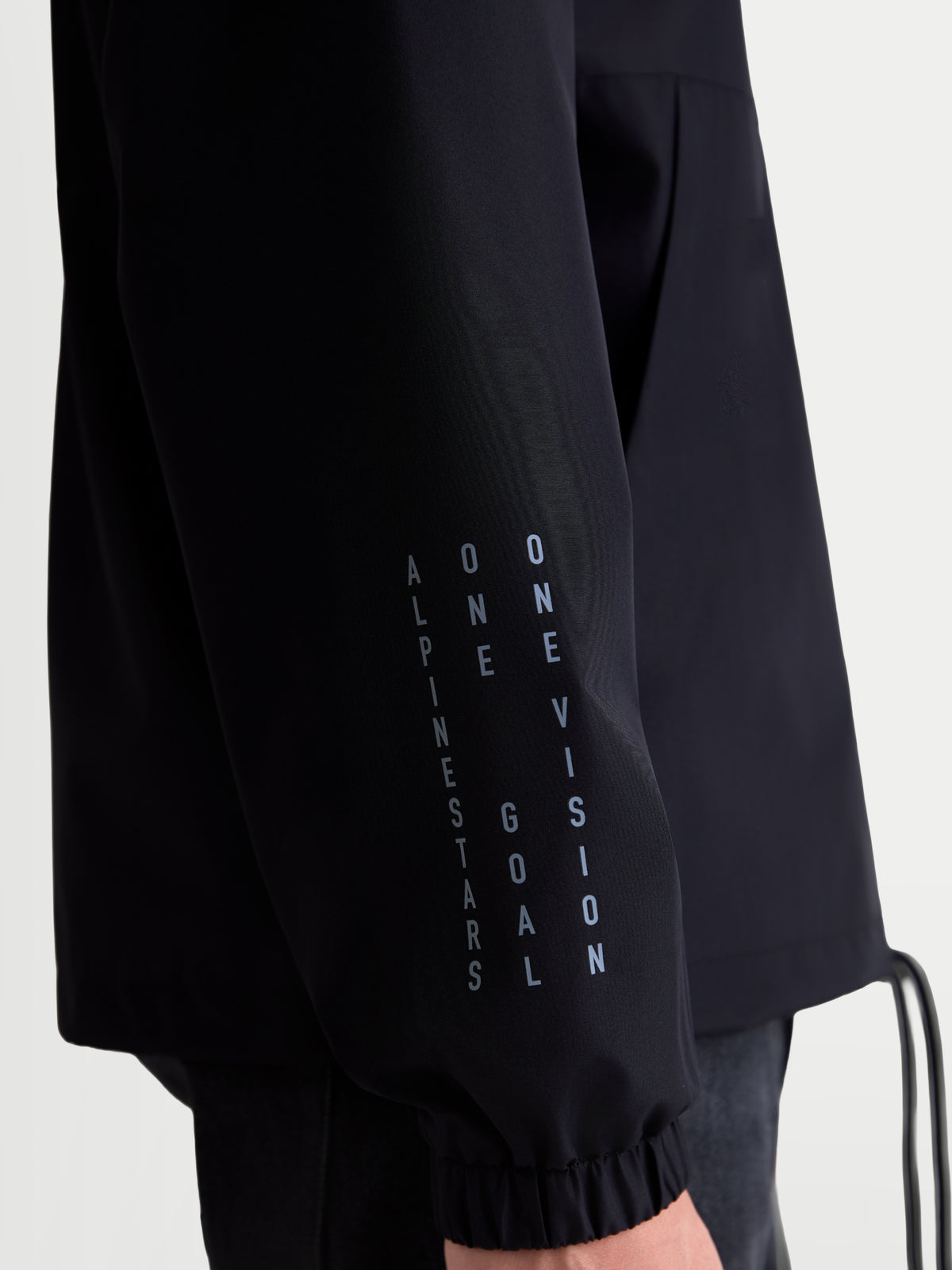 Alpinestars Speeder Resolve Winddichte Jacke, schwarze leichte Sportswear-Jacke, Nahaufnahme des Ärmels mit vertikalem Branding und elastischem Bündchen, winddichte Konstruktion aus 100 % Polyester für den urbanen Einsatz.