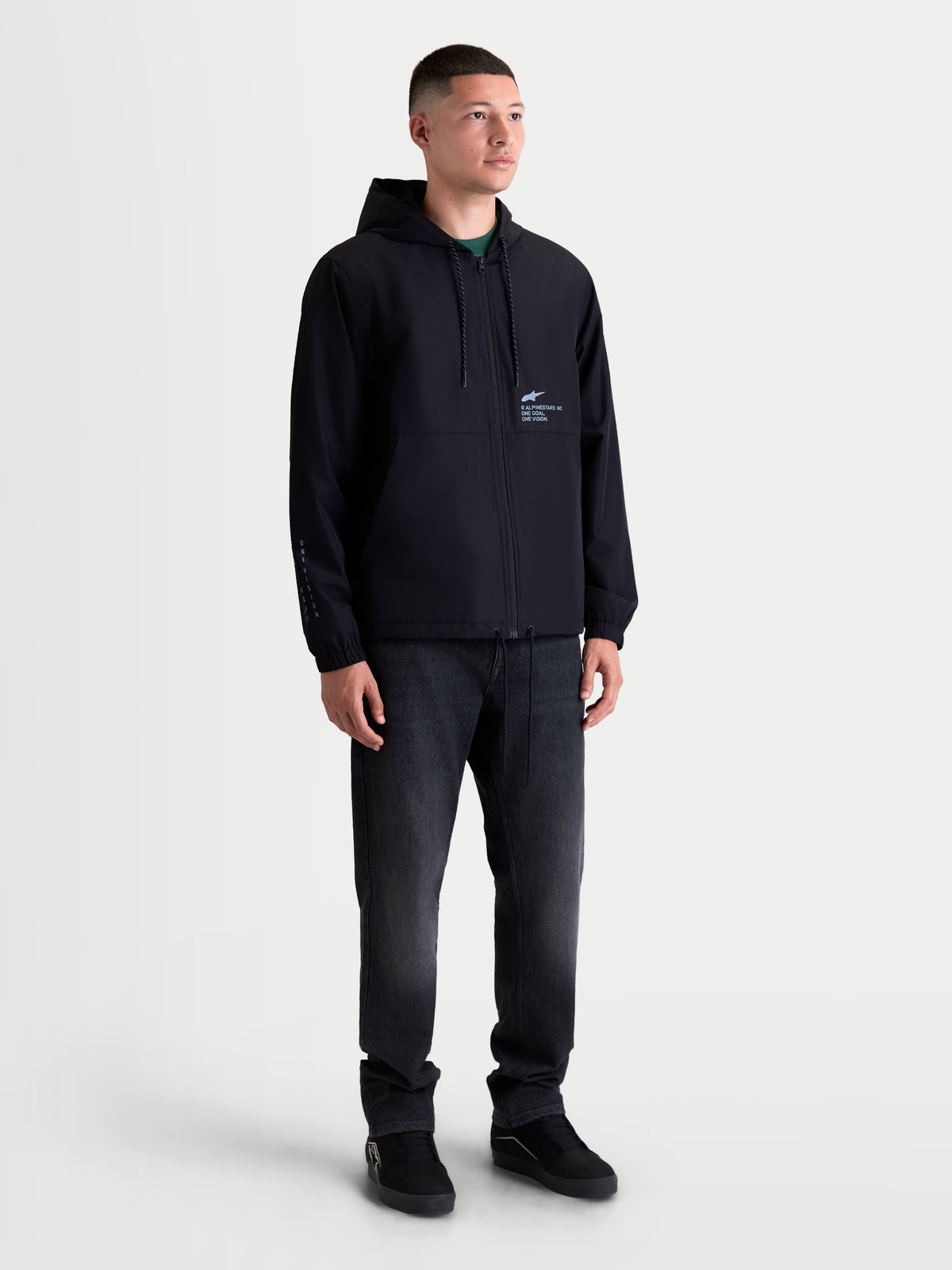 Alpinestars Speeder Resolve Winddichte Jacke, leichte Herren-Kapuzenjacke, Schwarz, mit netzgefütterter Innenseite, verstellbarer Kapuze mit Kordelzug und Alpinestars Motorsport-Branding auf der Brust und am Ärmel, am Model für urbanes Layering gezeigt.