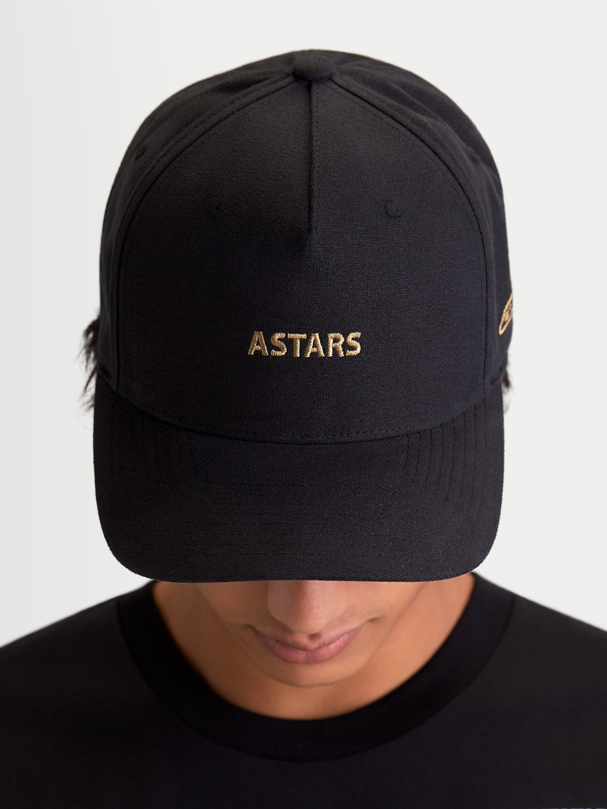 Alpinestars County Canvas Kappe, schwarze Sportswear-Kappe mit goldener ASTARS-Logo-Stickerei, gebogenem Schirm und strukturierter Krone, Unisex Motorsport-inspirierte Kappe aus Oxford-Peach-Gewebe, Frontansicht auf Modell.