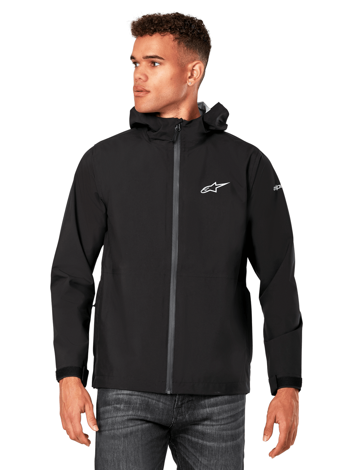 Male model wearing Alpinestars Kascade Rain Jacket, Black, white chest logo and sleeve branding, hooded rain shell with coated front zipper and velcro cuffs, paired with dark jeans, casual motorsport-inspired streetwear