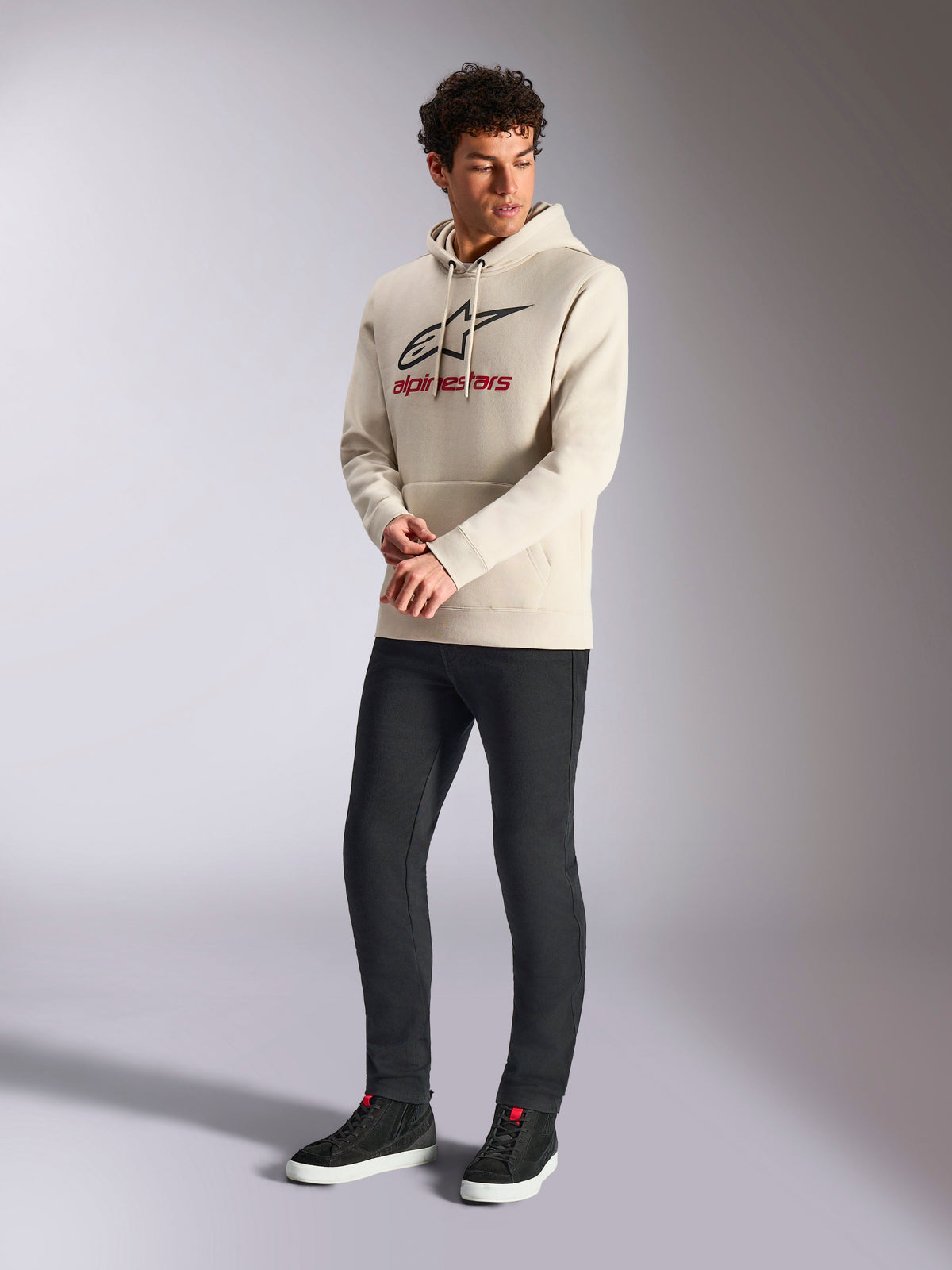 Alpinestars Always V3 Hoodie, cremeweißer Fleece-Kapuzenpullover, frontaler Logo-Print mit schwarzem Stern und rotem Branding, getragen von einem männlichen Model mit schwarzer Hose und schwarzen Sneakern, lässige Motorsport-Streetwear