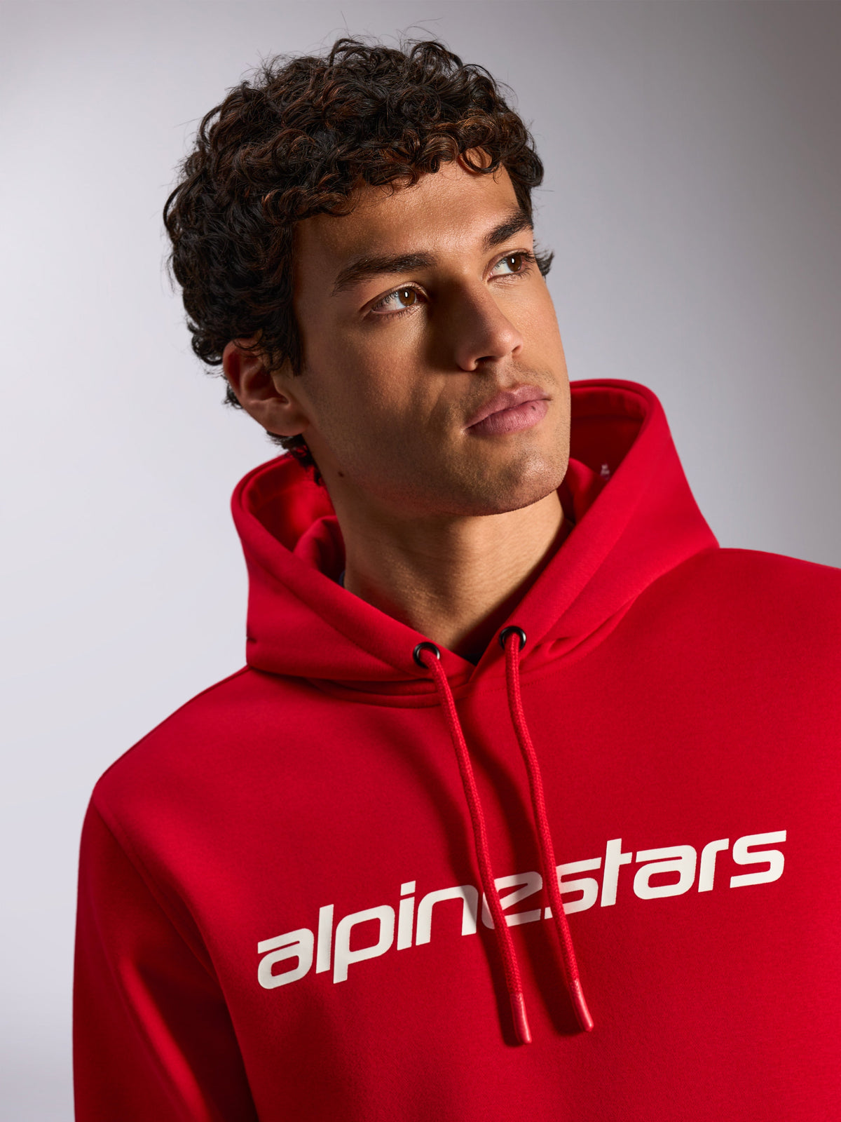 Alpinestars Linear Wordmark V3 Hoodie, roter Kapuzenpullover aus Fleece, weißer Alpinestars-Schriftzug auf der Brust, Kapuze mit Kordelzug und getauchten Spitzen, Lifestyle-Aufnahme eines männlichen Models