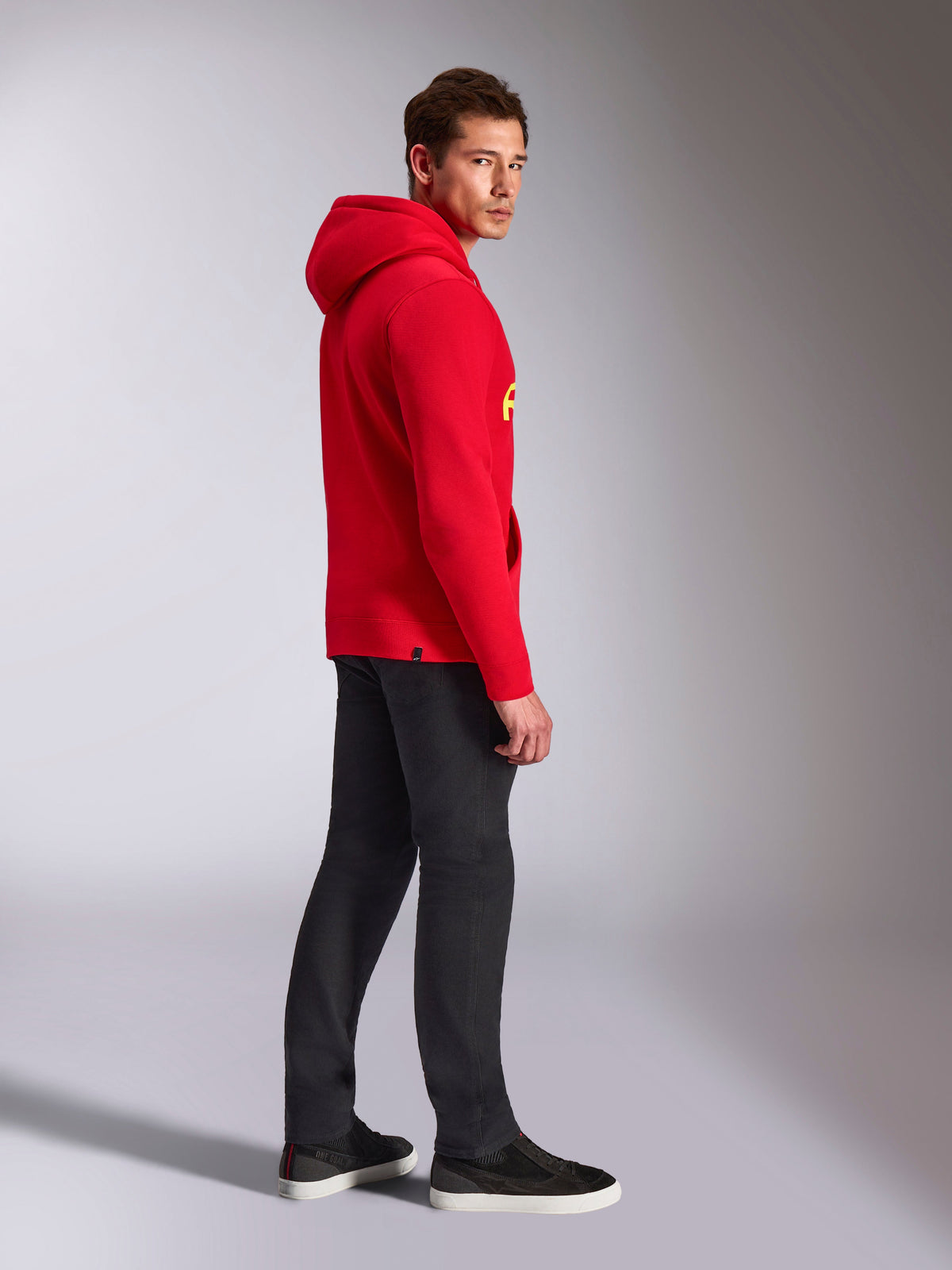 Alpinestars Ride V3 Hoodie, lässige Sportbekleidung, Rot mit Hi-Vis Gelbem Branding, männliches Model trägt roten Hoodie mit schwarzer Hose und Sneakern, Seitenprofil, Fleece-Pullover-Design mit Rippbündchen an Ärmeln und Saum.