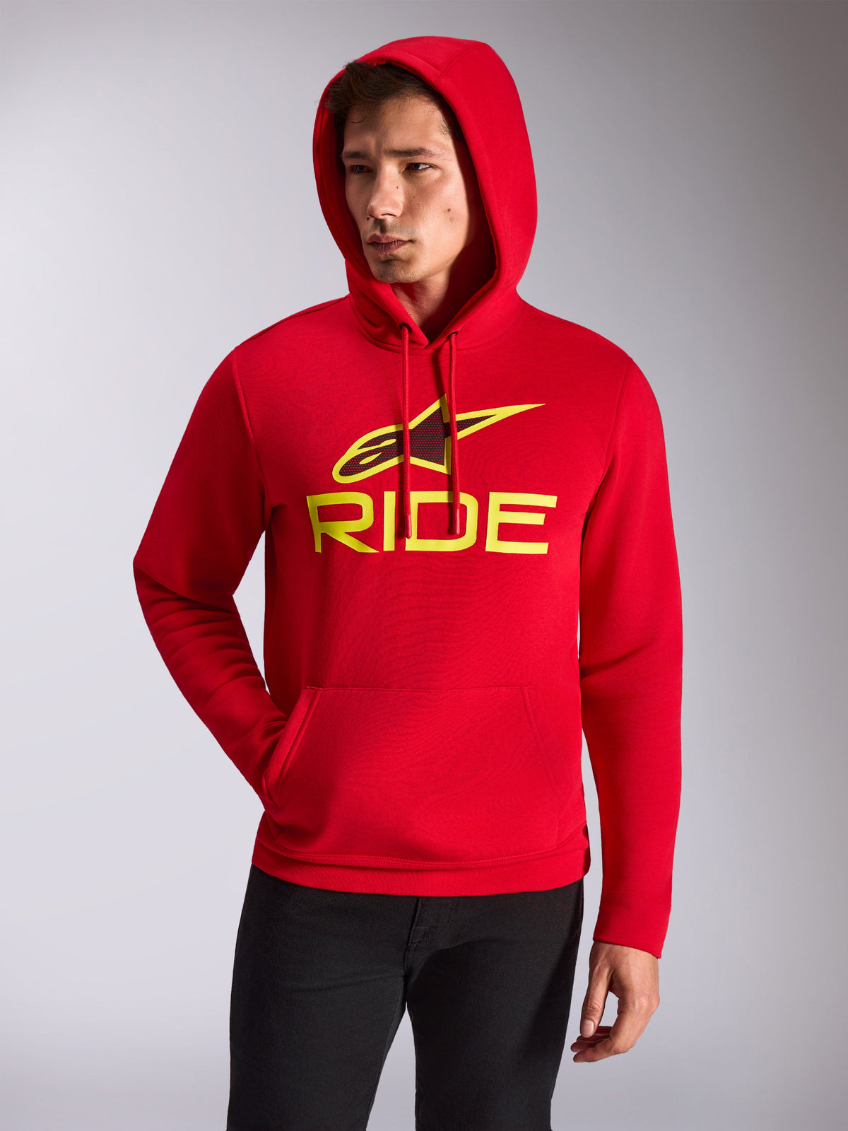 Alpinestars Ride V3 Hoodie, männliches Model trägt roten Kapuzenpullover aus Fleece, neongelbes RIDE-Logo und Alpinestars-Stern auf der Brust, lässige Motorsport-Streetwear, Kängurutasche vorne, getragen mit schwarzer Hose.