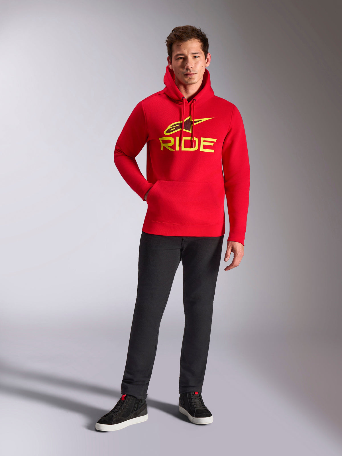 Alpinestars Ride V3 Hoodie, roter Fleece-Pullover, leuchtend gelbes RIDE-Logo mit Astars-Symbol auf der Brust, getragen mit schwarzer Hose und schwarzen High-Top-Sneakern, lässige, vom Motorsport inspirierte Streetwear