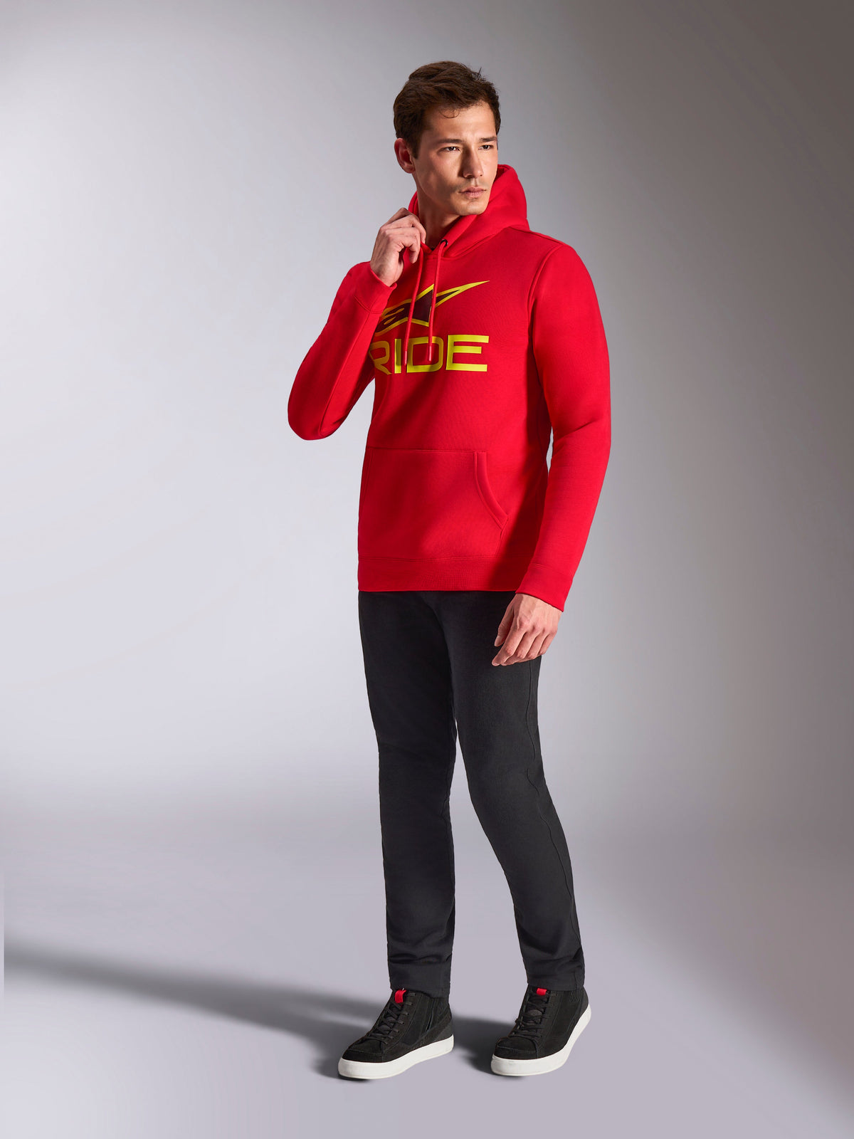 Alpinestars Ride V3 Hoodie, roter Kapuzenpullover, gelbes und schwarzes „RIDE“-Logo auf der Brust, getragen von einem Model mit schwarzer Hose und schwarzen Sneakern, lässige, vom Motorsport inspirierte Streetwear