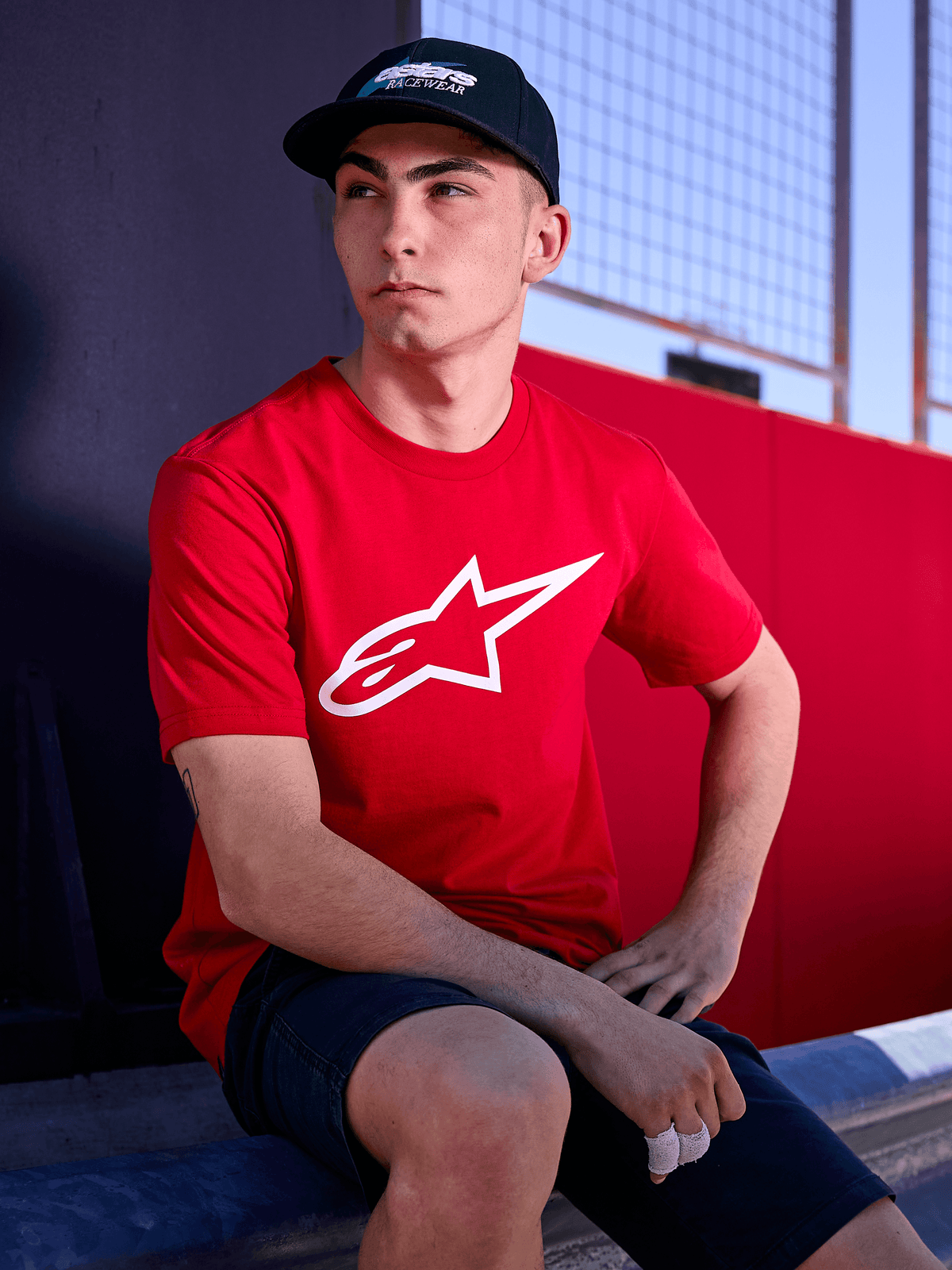 Alpinestars Profile Kappe in Schwarz, Lifestyle-Aufnahme eines Mannes mit der Kappe mit gebogenem Schirm, rotes T-Shirt mit weißem Alpinestars-Logo, lässige, vom Motorsport inspirierte Streetwear