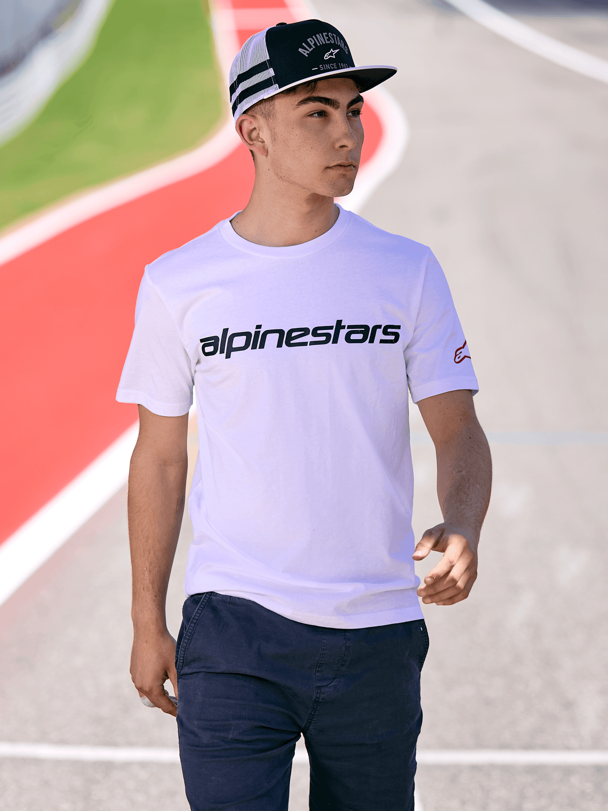 Man wearing Alpinestars Backline Trucker Hat in Black and White, paired with a white Alpinestars logo t-shirt and dark blue pants, casual motorsport streetwear on an outdoor track background