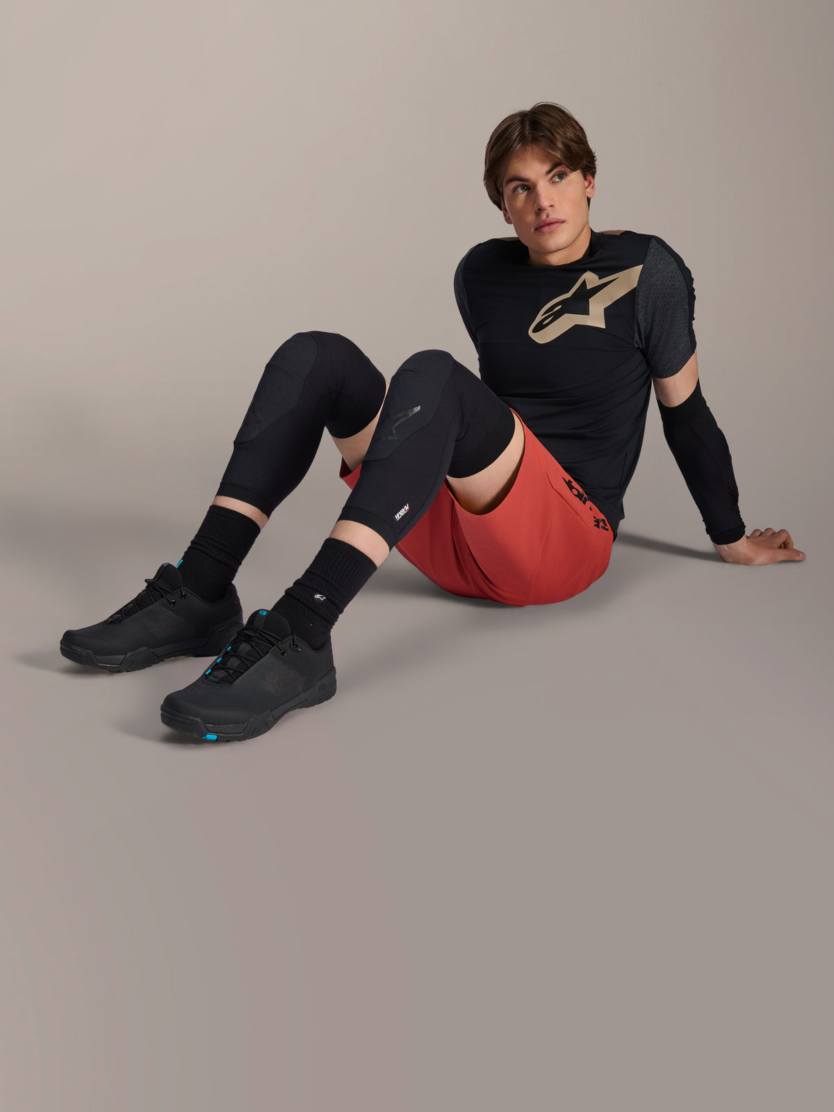 Alpinestars A-Motion Plasma Ellbogenärmel, getragen von einem männlichen Fahrer, der auf einem Studioboden sitzt, mit schwarzer Mesh-Konstruktion, kombiniert mit roten Shorts und einem schwarzen Alpinestars-Trikot mit hellbraunem Logo, Performance-Mountainbike-Schutz für Trail-Riding.