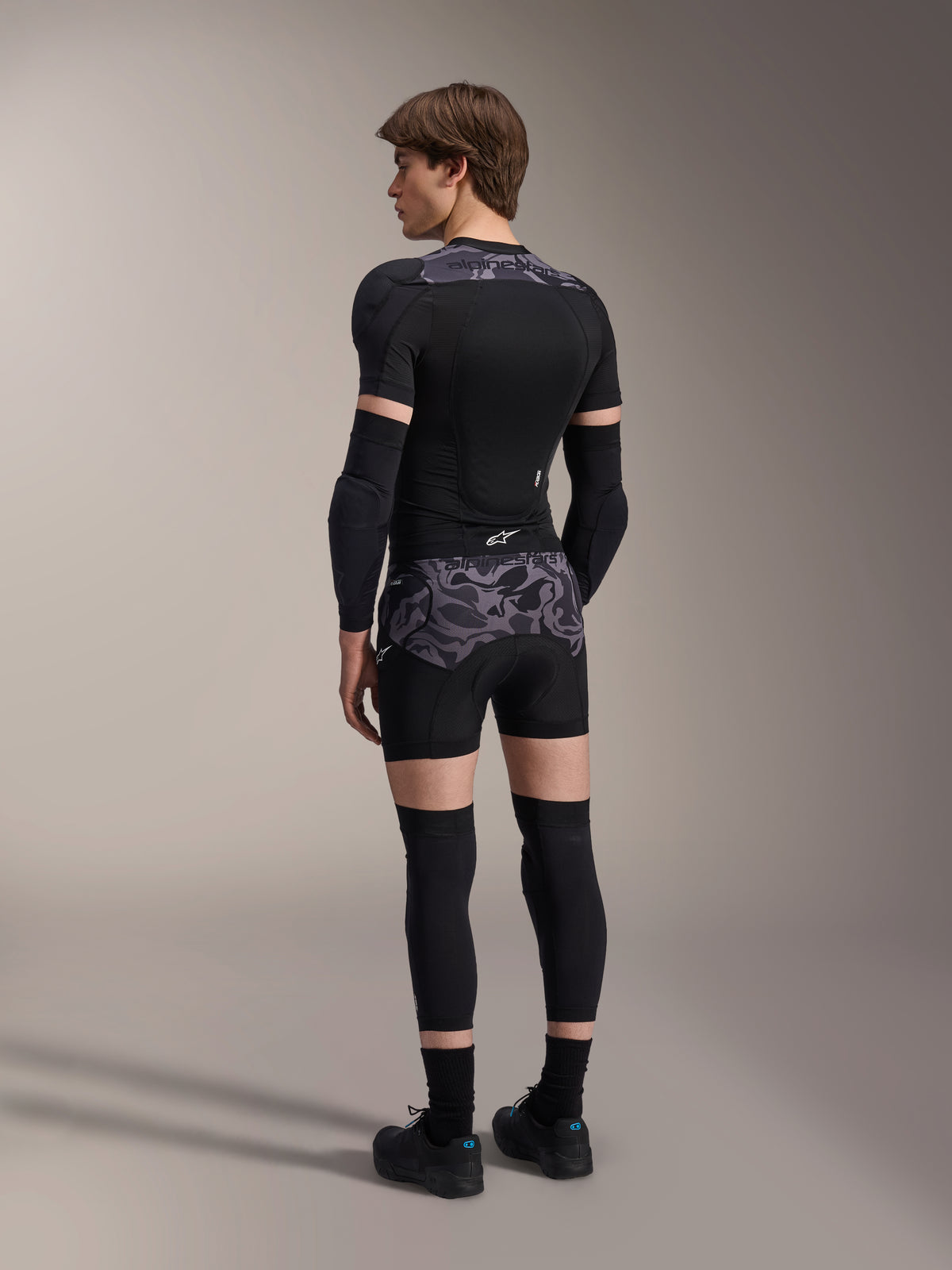 Modell trägt Alpinestars A-Motion Plasma Protektorenshorts in Black Camo und passende A-Motion Plasma Protektorenjacke, Rückansicht mit grauem Camouflage-Muster, Ellbogenprotektoren, Knieprotektoren und Radschuhe, komplette Mountainbike-Schutzausrüstung.