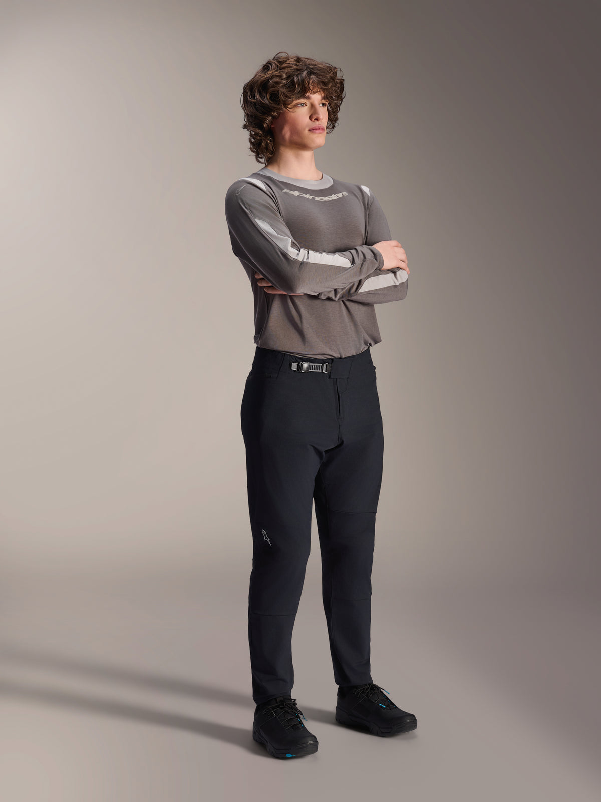 Männliches Model in Alpinestars A-Dura Elite Hose, schwarze MTB-Hose, kombiniert mit einem grauen Langarmtrikot und schwarzen Radschuhen, Ratschenverschlusssystem vorne, professionelle Mountainbike-Bekleidung für Trail- und Enduro-Fahrten