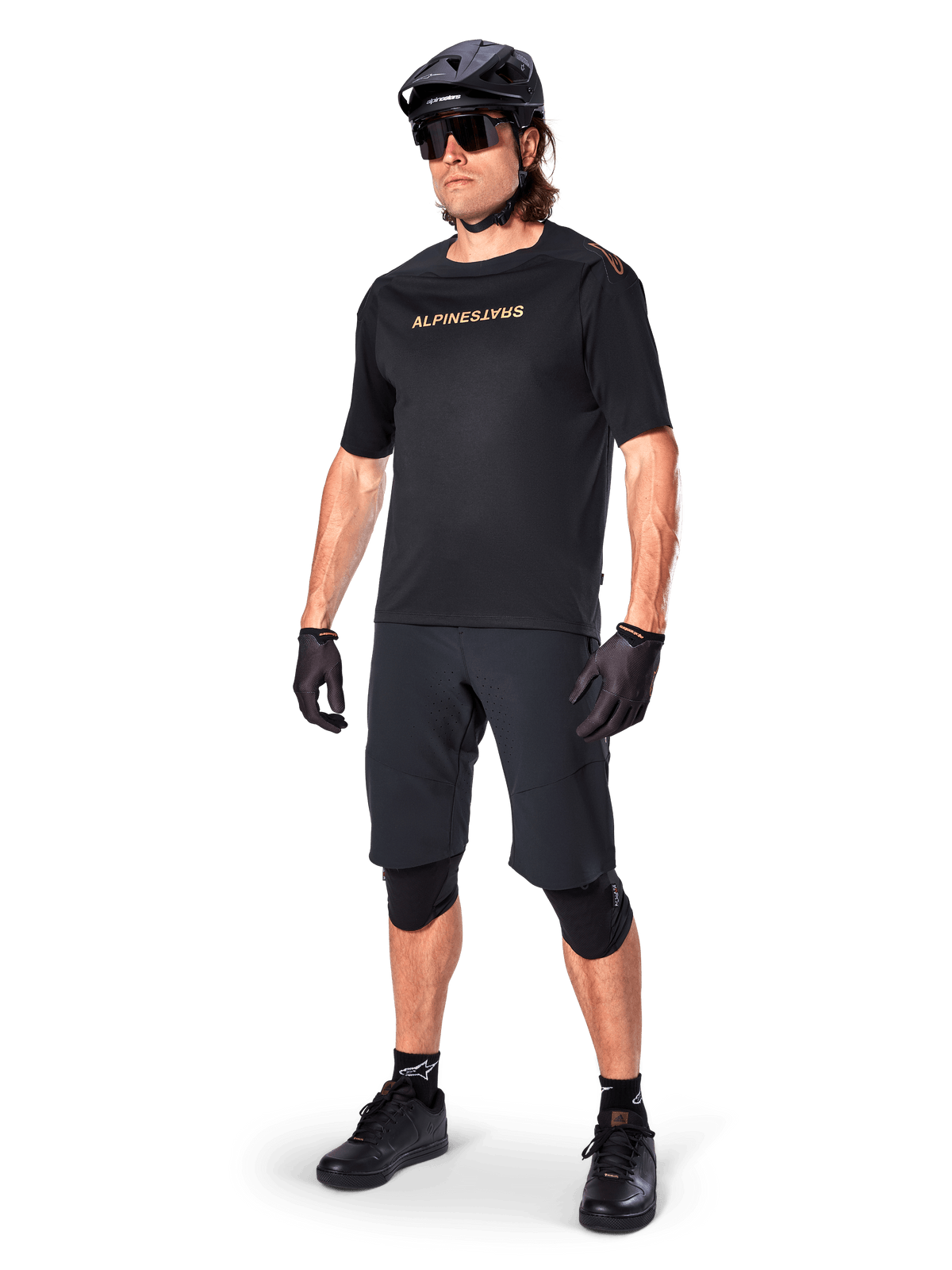 Radfahrer in Alpinestars A-Aria Elite Shorts, schwarze Mountainbike-Shorts, kombiniert mit einem passenden schwarzen Trikot mit goldenem Branding, schwarzer Helm, Sonnenbrille und Knieschützer, komplettes Performance-Outfit für den Offroad-Radsport