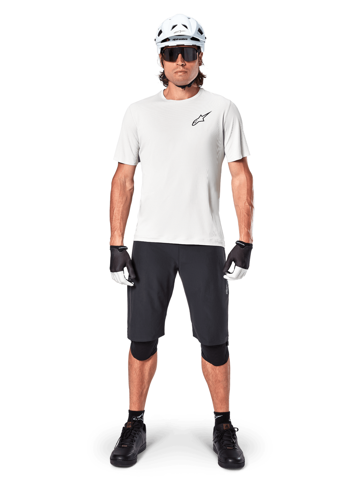 Männlicher Mountainbiker in Alpinestars A-Aria Shorts, Schwarz, kombiniert mit einem weißen Performance-Trikot, weißem Helm und schwarzen Knieprotektoren, professionelle Offroad-Radsportausrüstung in einer Ganzkörper-Frontansicht