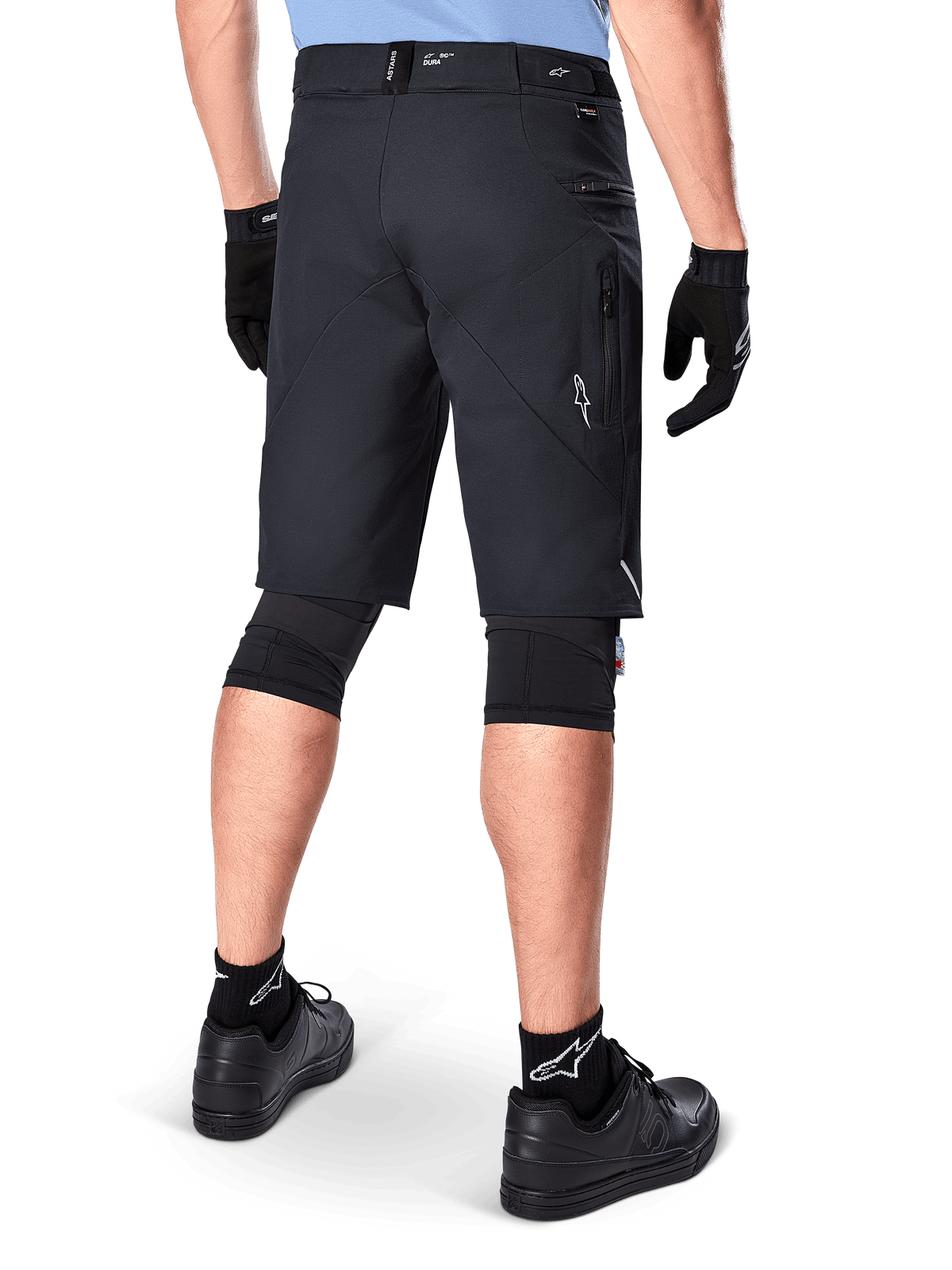 Alpinestars A-Dura Elite Shorts, Schwarz, Rückansicht eines Fahrers in Mountainbike-Ausrüstung einschließlich schwarzer Knieschützer, Handschuhe und Socken mit weißen Logos, mit seitlichen Reißverschlusstaschen und reflektierenden Akzenten auf strapazierfähigem Cordura-Gewebe.