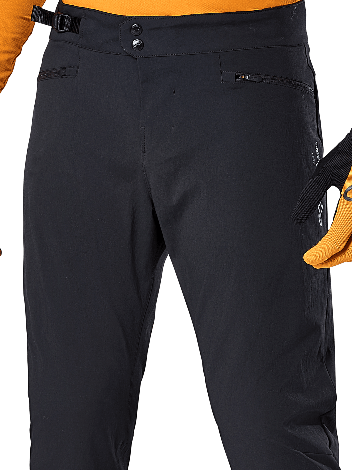 Alpinestars A-Dura Hose, schwarze MTB-Radhose, Detailansicht mit doppeltem Druckknopfverschluss, verstellbarem seitlichem Taillenversteller und Reißverschlusstaschen, kombiniert mit einem orangefarbenen Trikot und Handschuhen