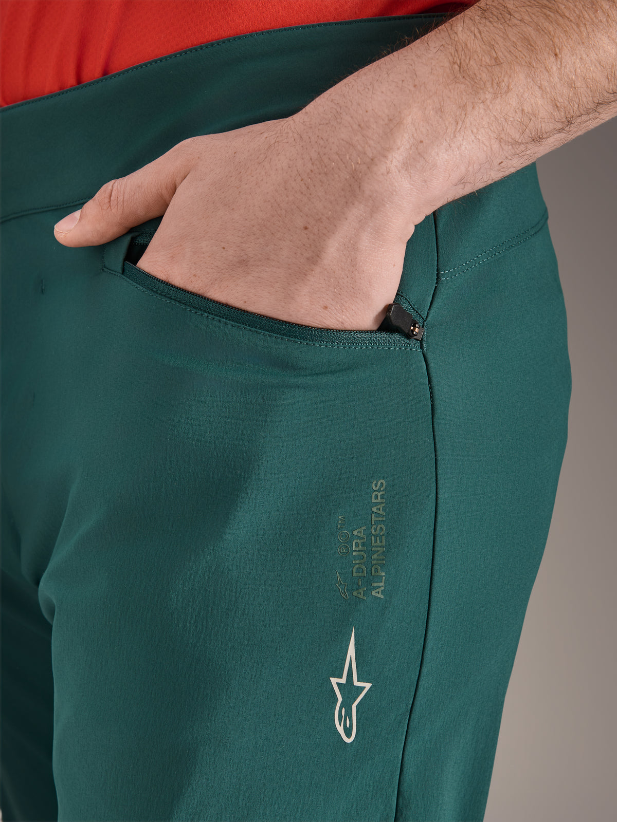 Alpinestars A-Dura Hose in Dark Pine Grün, Nahaufnahme einer Reißverschlusstasche mit Hand, weißes Alpinestars-Logo und Markenschriftzug am Oberschenkel, strapazierfähige MTB-Fahrradhose