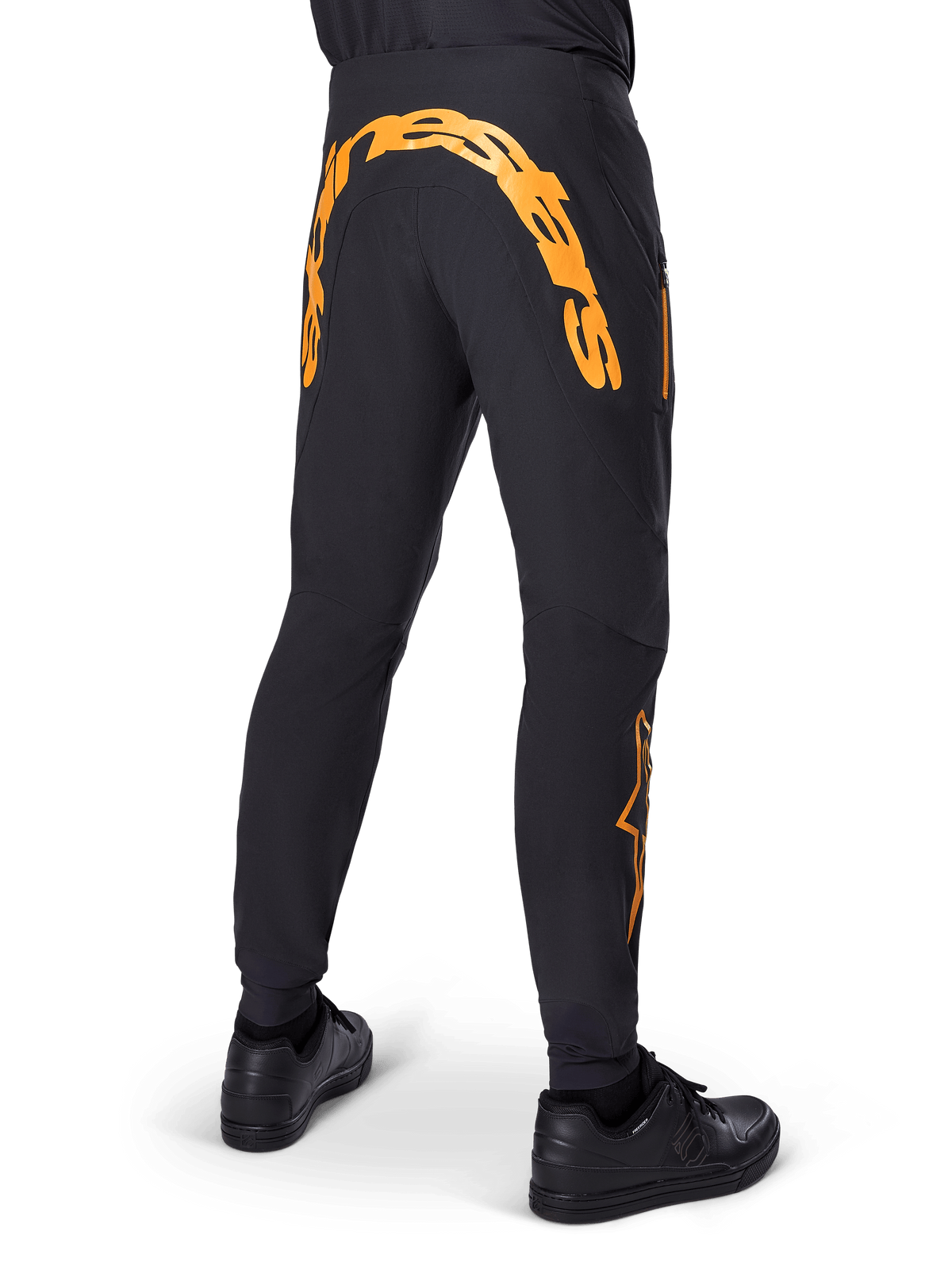 Alpinestars A-Supra Race Pants, schwarze MTB-Hose, Rückansicht mit großem orangefarbenem Alpinestars-Schriftzug über der hinteren Passe, orangefarbenes Logo am Unterschenkel, Performance-Passform mit Reißverschlusstasche, getragen mit schwarzen Schuhen