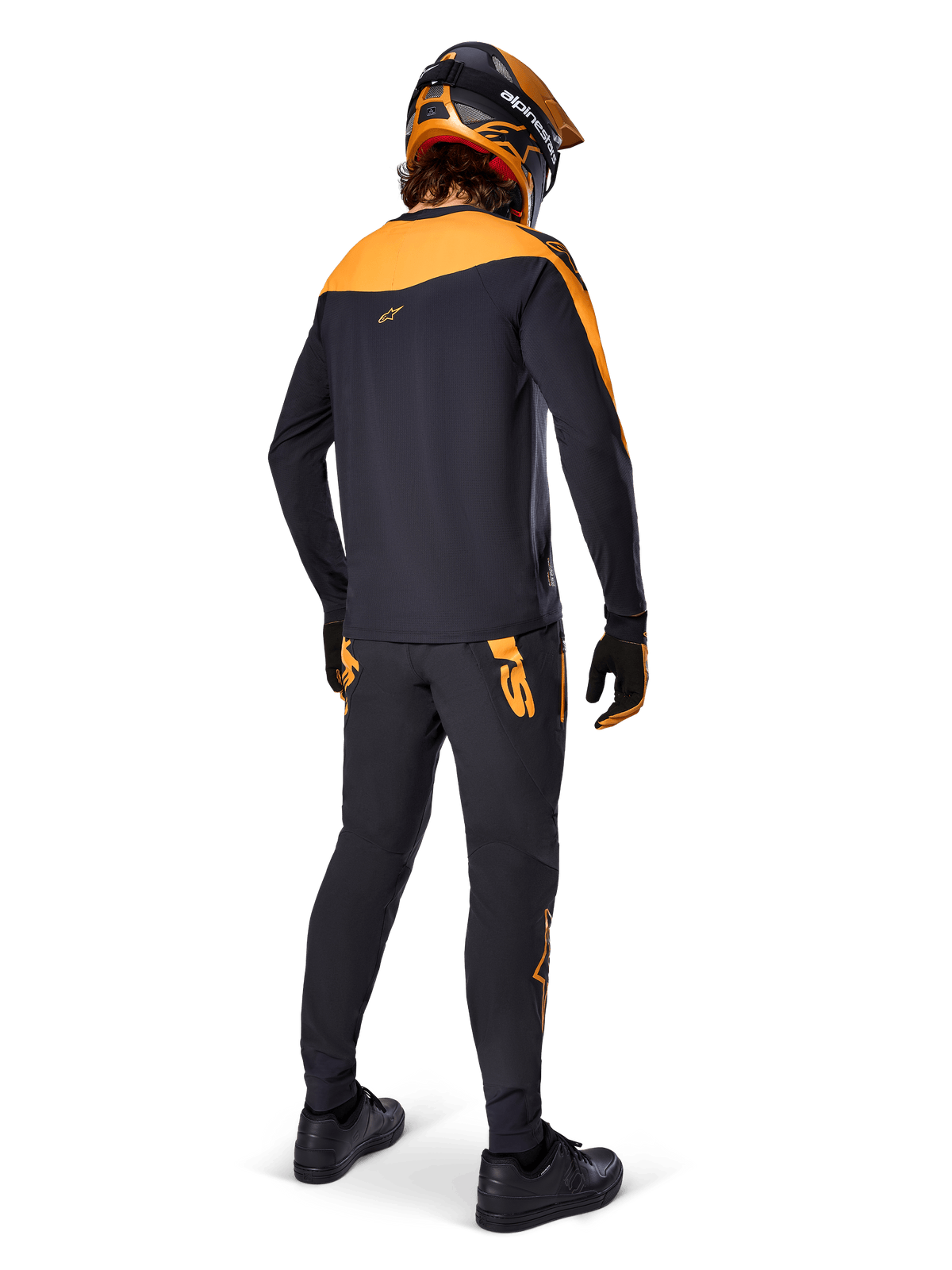 Alpinestars A-Supra Race Pants, MTB-Renn-Hose, Schwarz mit orangefarbenen Logos, Rückansicht eines Fahrers in passendem schwarz-orangefarbenem Trikot, Helm und Handschuhen, hebt den MX-inspirierten Schriftzug auf der Rückseite und die Performance-Passform für technische Trails hervor.