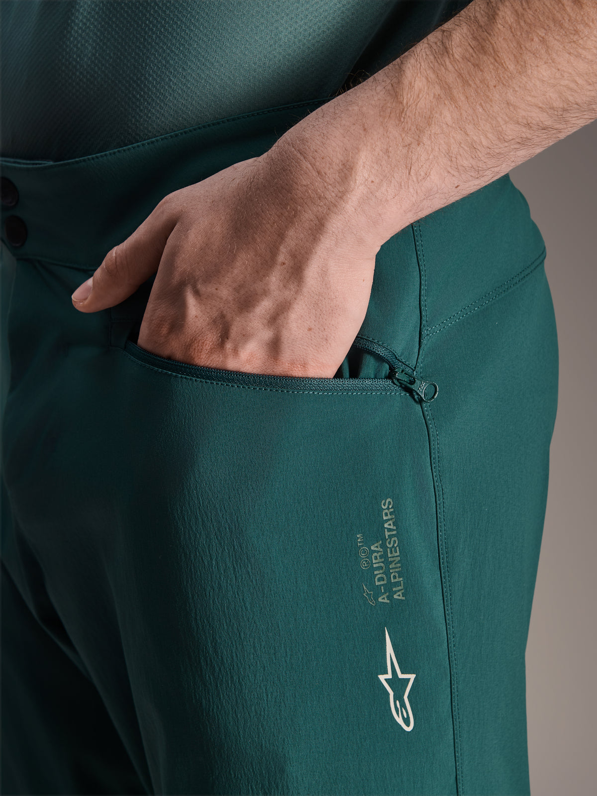 Alpinestars A-Dura Shorts in Dark Pine, Nahaufnahme einer Hand in der Reißverschlusstasche mit Mesh-Futter, weißes Alpinestars Stern-Logo und A-Dura Branding, strapazierfähige Textilkonstruktion für Mountainbiking