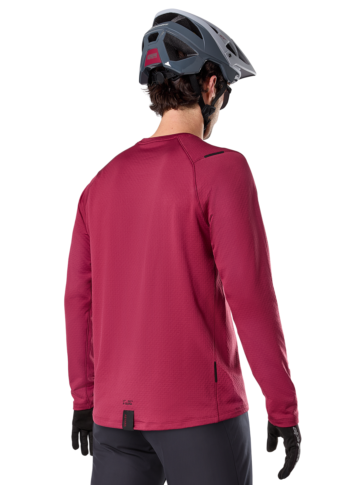 Alpinestars A-Dura Thermal Trikot, Langarm-Mountainbike-Trikot, Burgund, Rückansicht am Model mit grauem MTB-Helm und schwarzen Handschuhen, mit verlängertem Rückenteil und kleinen Logo-Details, entwickelt für Trail-Fahrten bei kaltem Wetter.