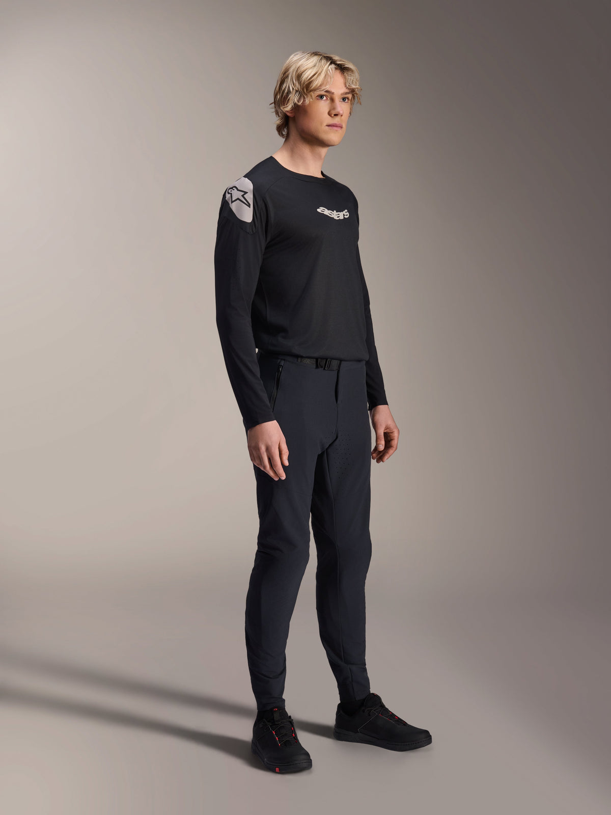 Model im Alpinestars A-Aria Polartec Elite Langarm-Trikot in Schwarz, kombiniert mit passender schwarzer technischer Mountainbike-Hose und schwarzen Schuhen, Performance-Radsportbekleidung für Offroad-Fahrten, Studioaufnahme.