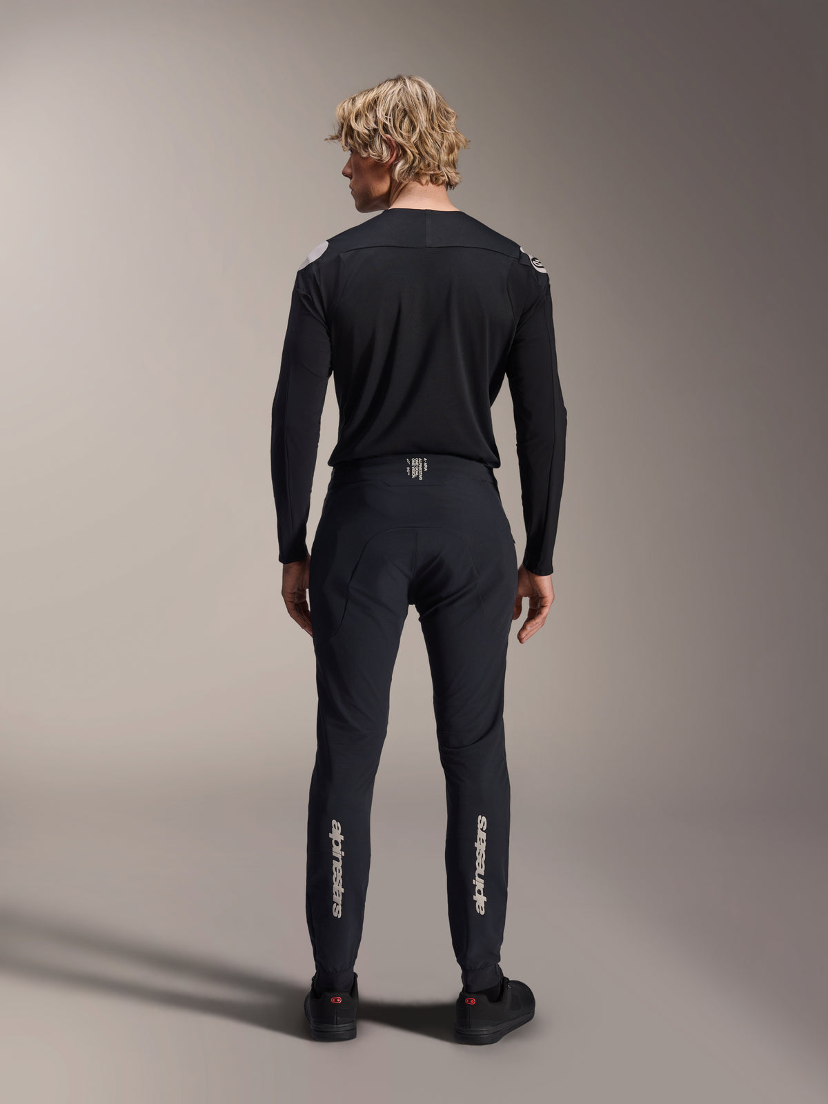 Alpinestars A-Aria Elite Hose, Black Titanium, Rückansicht an männlichem Model, mit silbernem Alpinestars-Branding an den Unterschenkeln, technische MTB-Hose mit Performance-Passform und feuchtigkeitsableitendem Detail am Bund.