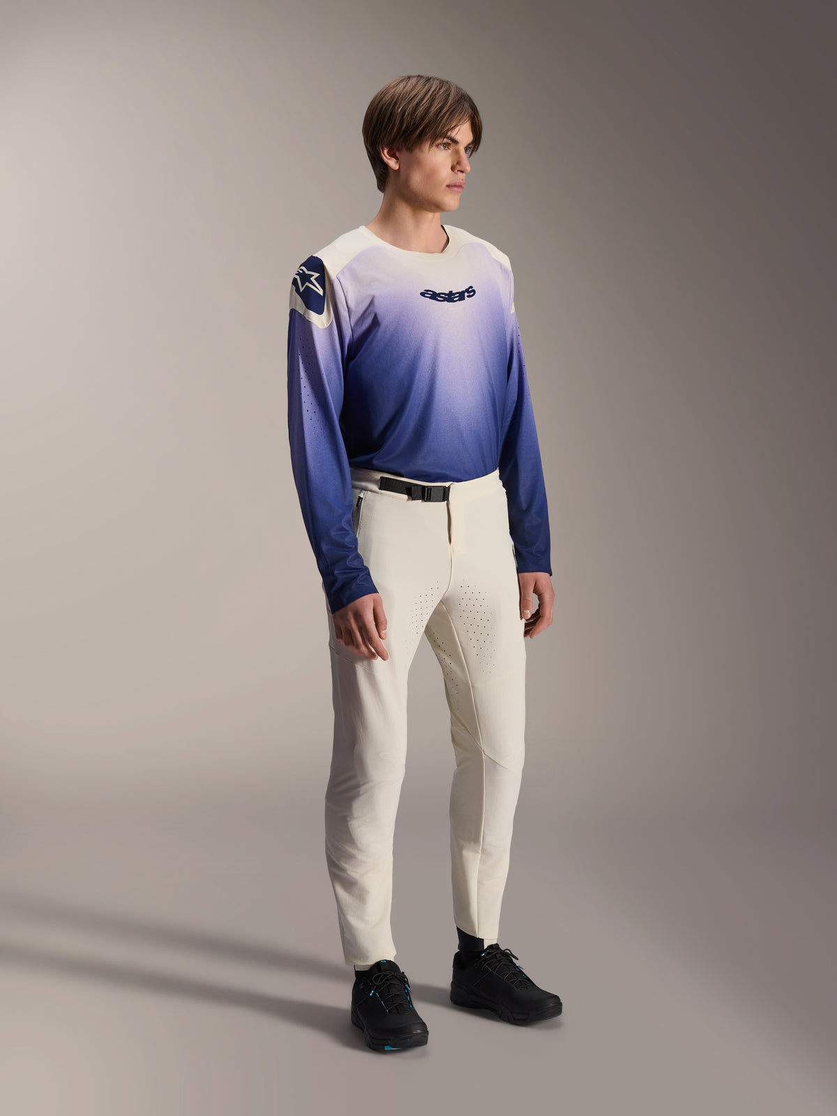 Model trägt die Alpinestars A-Aria Elite Hose in Ecru, eine naturweiße Mountainbike-Hose mit lasergeschnittener Belüftung und schwarzer Gürtelschnalle, kombiniert mit einem langärmeligen Trikot mit blau-violettem Farbverlauf, leistungsorientierte Fahrradausrüstung für Offroad-Trails