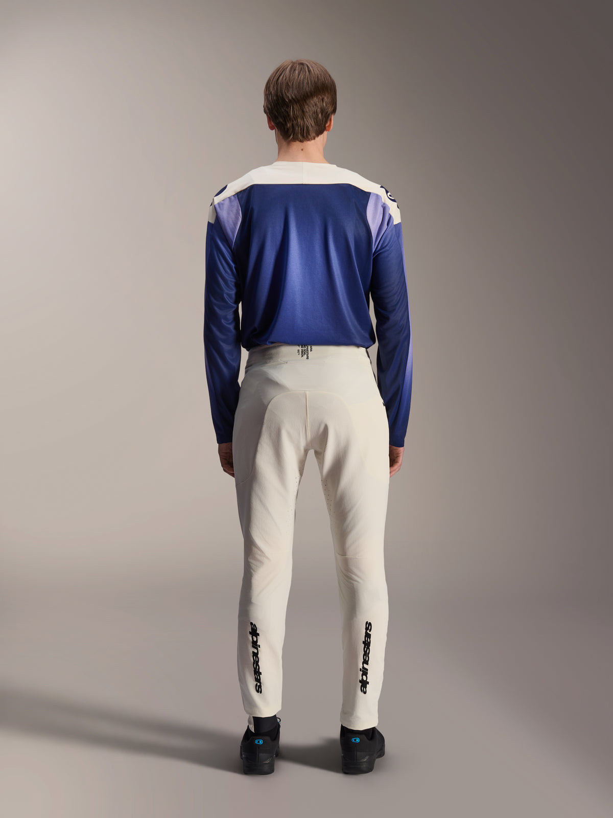 Alpinestars A-Aria Elite Hose in Ecru, Rückansicht an einem Model kombiniert mit einem blauen A-Aria Polartec Maker Elite Langarm-Trikot, naturweiße technische MTB-Hose mit schwarzen Logos an den Unterschenkeln, Performance-Radsportbekleidung für Mountainbiking