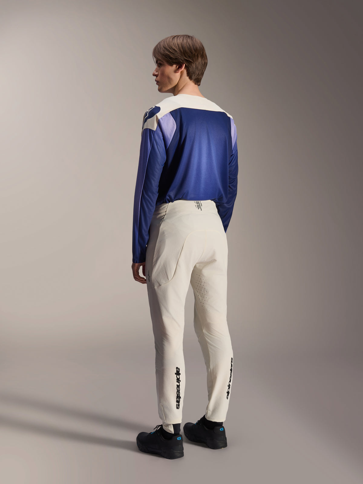 Alpinestars A-Aria Elite Hose, MTB-Hose, Ecru, Rückansicht an einem Model, mit schwarzem Schriftzug-Branding an den Unterschenkeln und Laserperforationen zur Belüftung, kombiniert mit einem blau-violetten Langarmtrikot und schwarzen Schuhen, professionelle Mountainbike-Ausrüstung