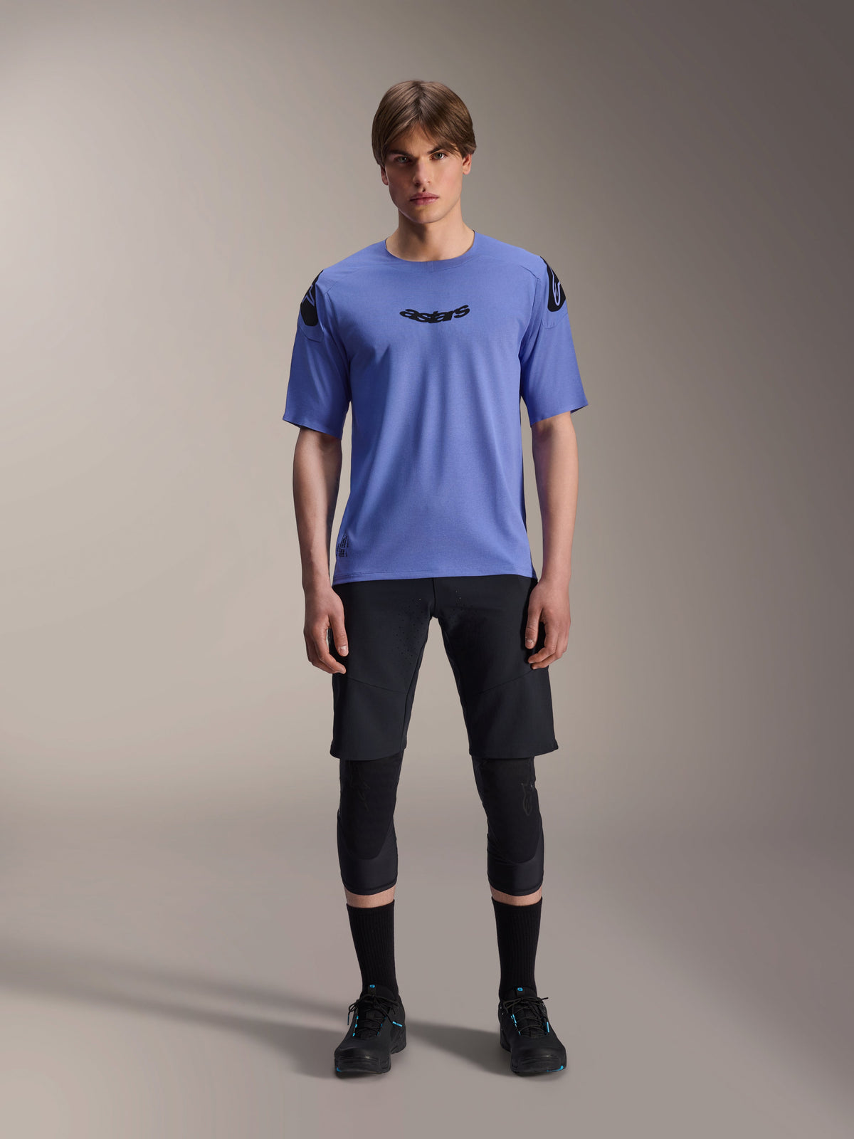 Männliches Model im Alpinestars A-Aria Polartec Elite Kurzarmtrikot in Baja Blue, schwarzes Astars-Logo auf der Brust, kombiniert mit schwarzen MTB-Shorts, Knieschützern und schwarzen Radschuhen, Studioaufnahme für Mountainbike-Bekleidung