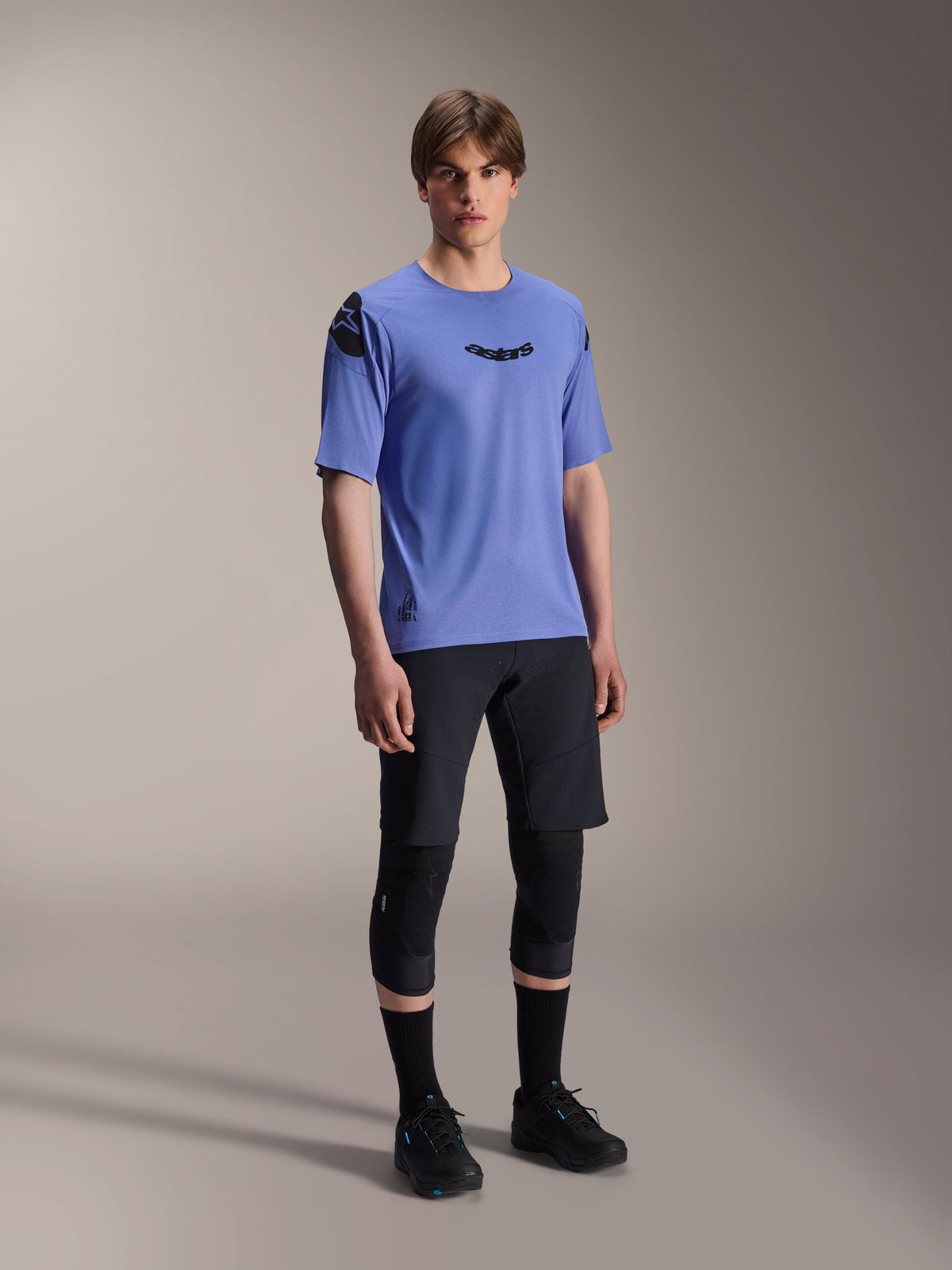 Männliches Model im Alpinestars A-Aria Polartec Elite Kurzarmtrikot in Baja Blue, kombiniert mit schwarzen MTB-Shorts und Knieschonern, mit schwarzen Astars-Logos auf der Brust und Schulter, hochwertige Performance-Mountainbike-Ausrüstung