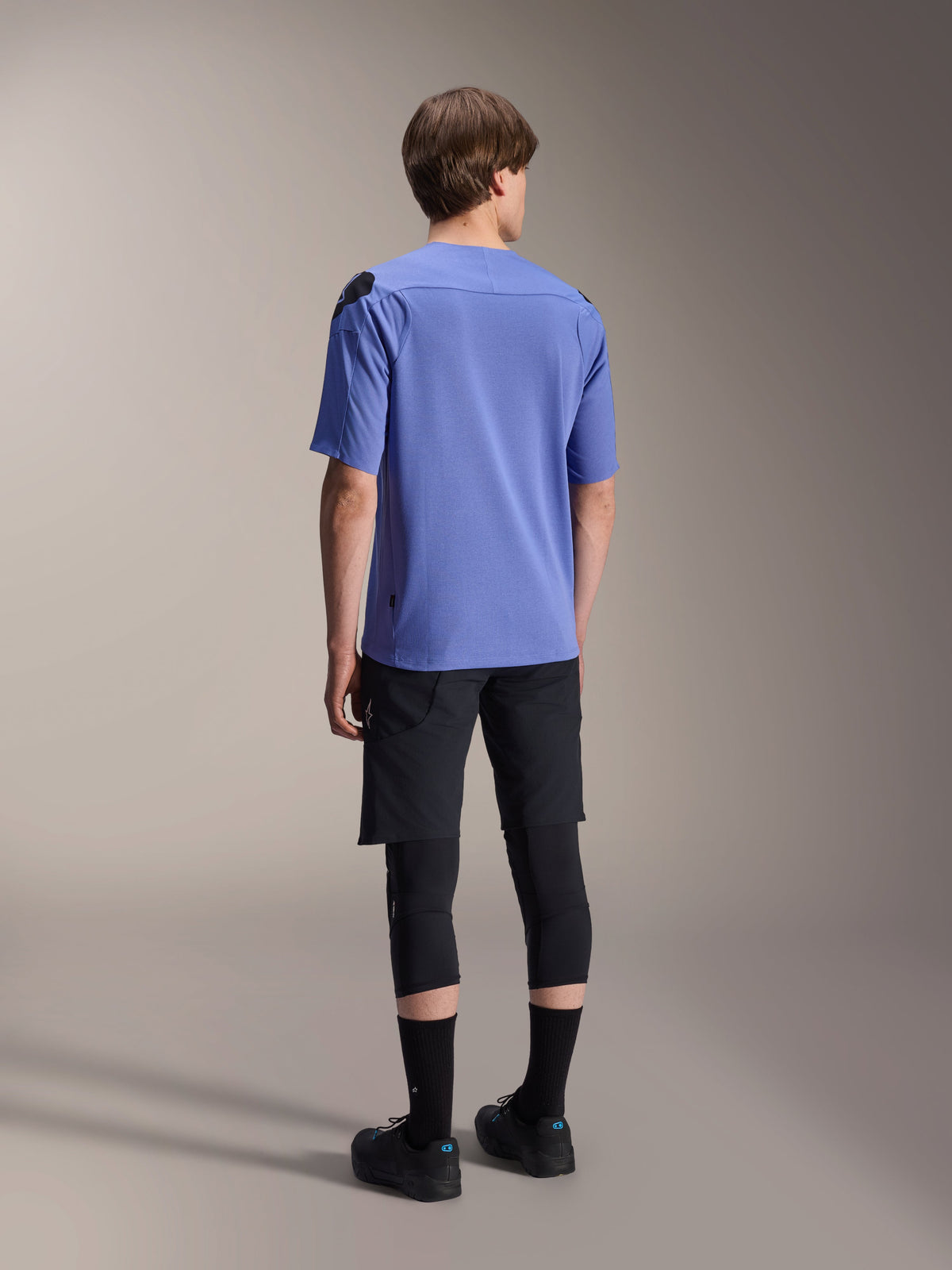 Alpinestars A-Aria Polartec Elite Kurzarmtrikot in Baja Blue, Rückansicht an einem männlichen Model, kombiniert mit schwarzen MTB-Shorts und Knieschutz, mit einer Performance-Passform und verlängertem Rückensaum für Trailfahrten.