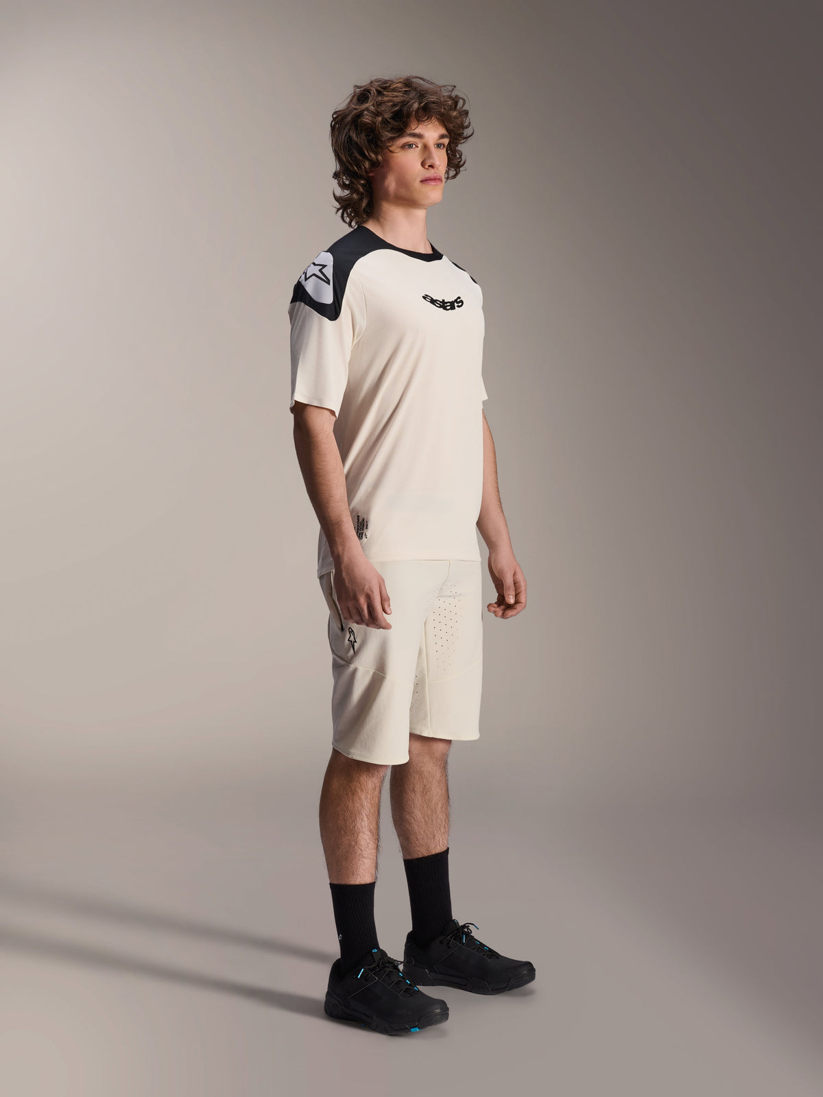 Alpinestars A-Aria Polartec Elite Kurzarmtrikot, Ecru mit schwarzen Schultereinsätzen und Logo, getragen von einem Model mit passenden Shorts und schwarzen Schuhen, Performance-Mountainbike-Bekleidung für Offroad-Trailfahrten.