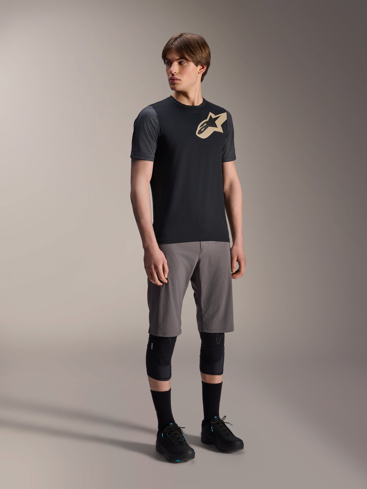 Male model wearing Alpinestars A-Dura Lipan Short Sleeve Jersey in Black, featuring dark grey sleeves and a tan Astars logo on the chest, paired with grey mountain bike shorts, black knee pads, and black cycling shoes, coordinated mountain bike riding gear