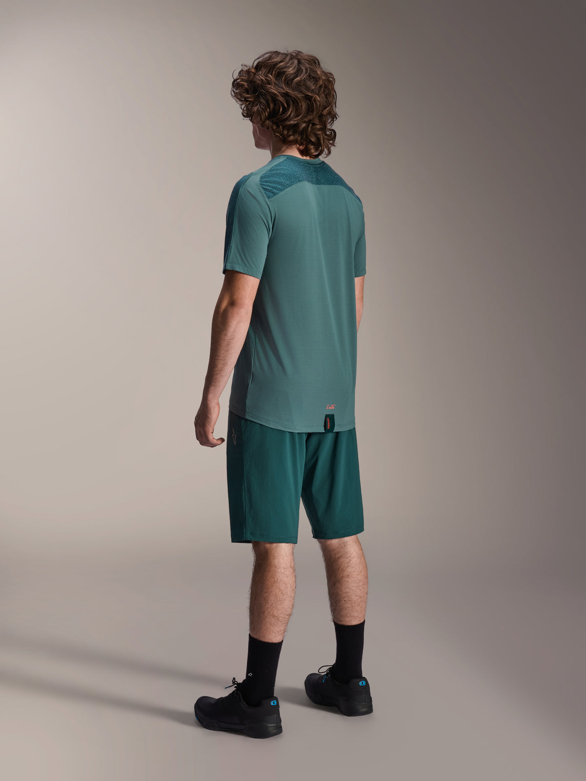 Alpinestars A-Dura Shorts in Dark Pine und passendes grünes Lipan Kurzarmtrikot, Rückansicht, kombiniert mit schwarzen Socken und schwarzen Mountainbike-Schuhen, professionelle MTB-Fahrausrüstung