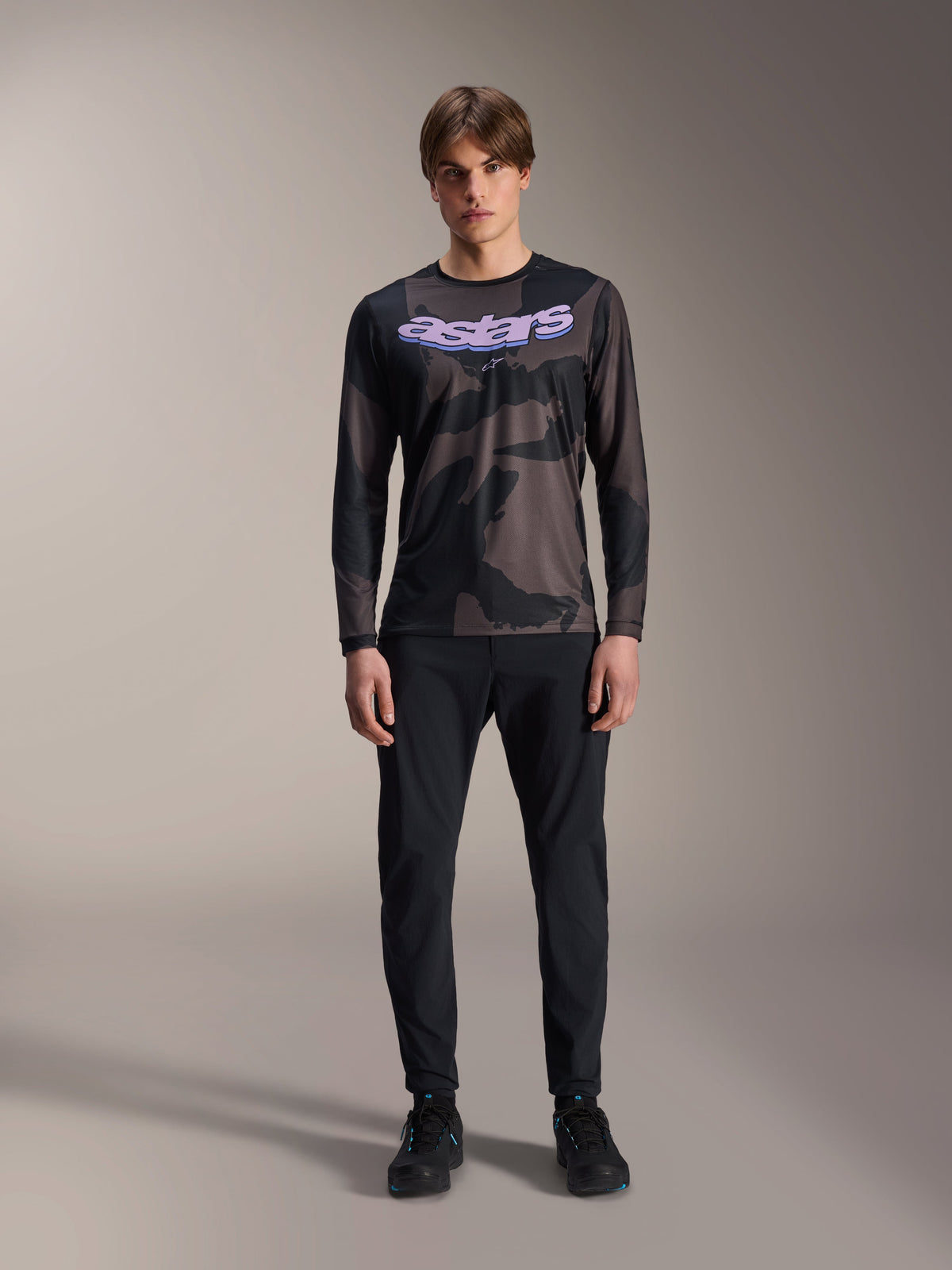 Modell im Alpinestars A-Dura Hollow Camo Langarm-Trikot in Black Camo, mit lila „astars“-Logo auf der Brust, kombiniert mit schwarzer Hose und schwarzen Radschuhen, Mountainbike-Ausrüstung